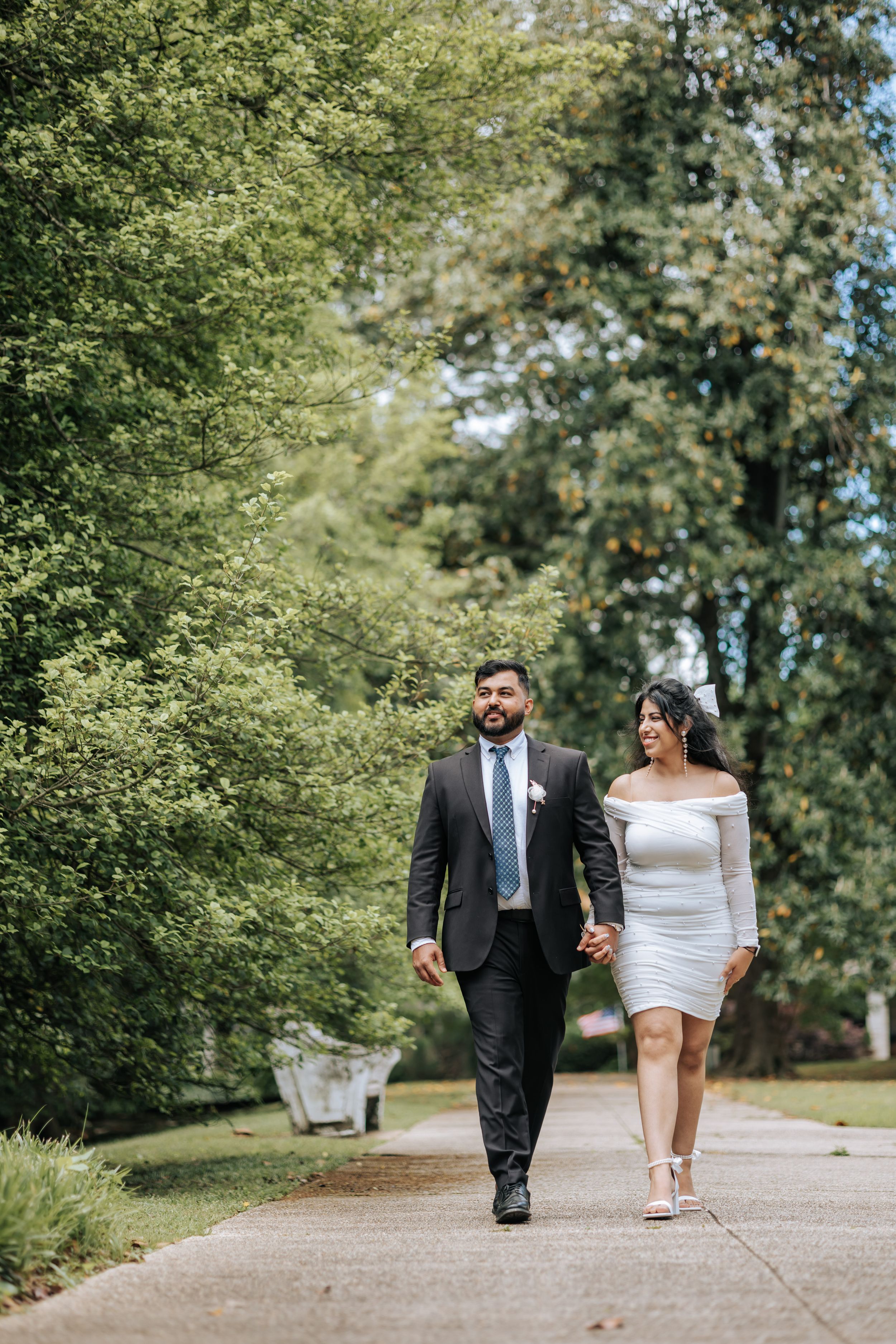 Couple in wedding attire walking hand-in-hand on a tree-lined path, man in dark suit, woman in white off-shoulder dress.