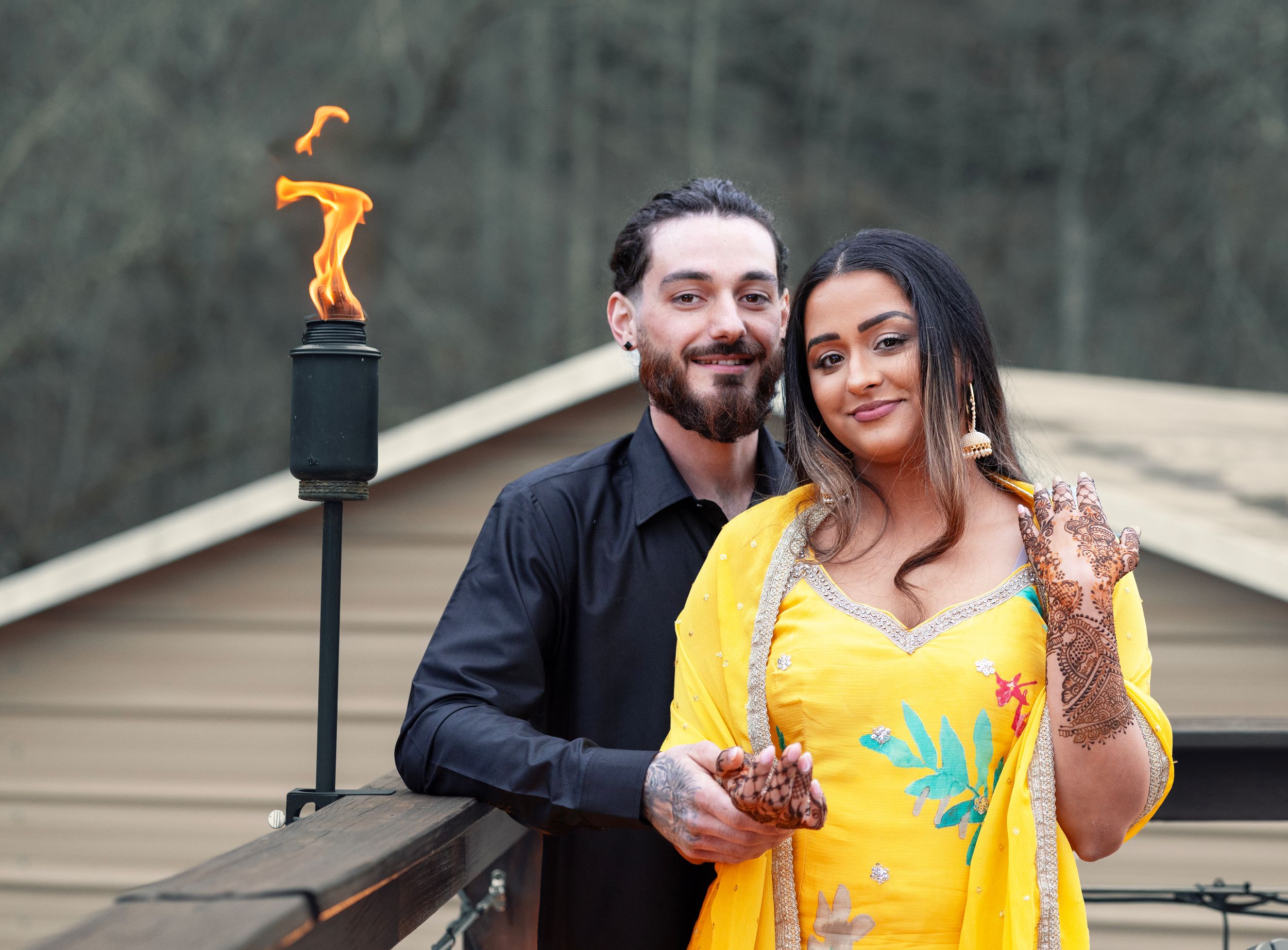 Couple in traditional South Asian attire posing outdoors near a lit tiki torch, woman wearing yellow saree with henna.