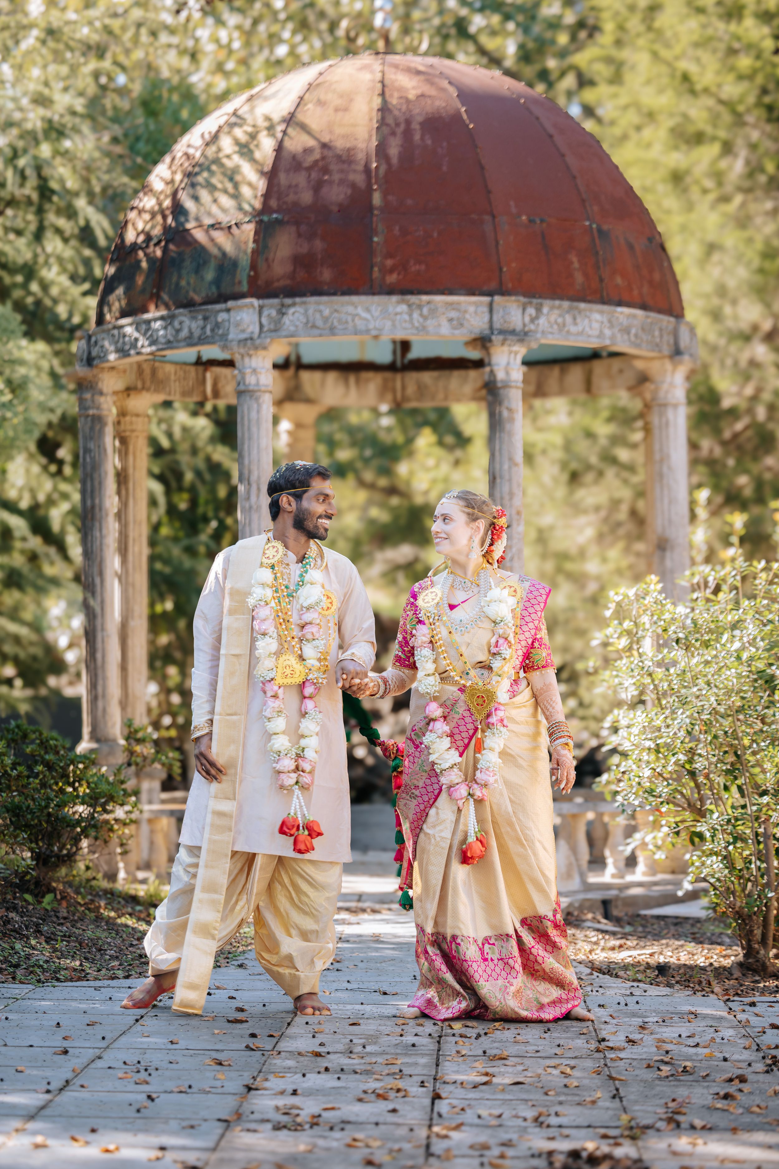 Indian groom in cream sherwani and bride in gold saree exchange smiles during traditional wedding ceremony in garden gazebo.