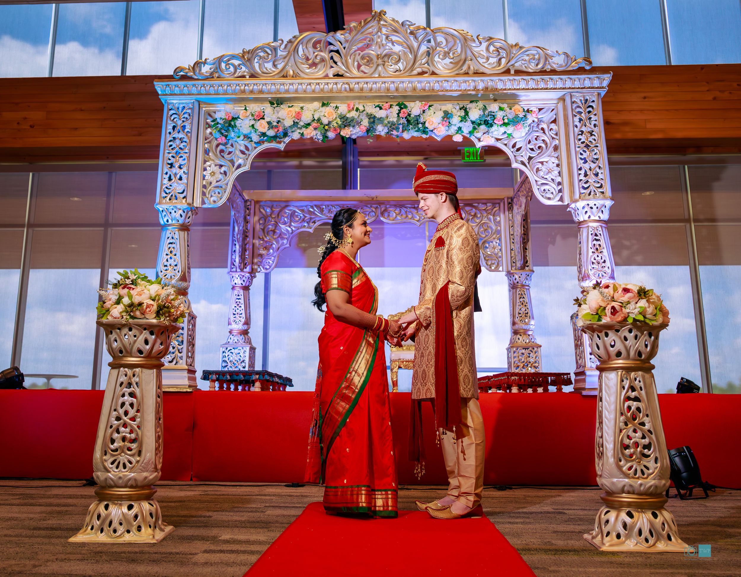 Couple in traditional Indian wedding attire holding hands under ornate white mandap with red carpet and floral decorations.