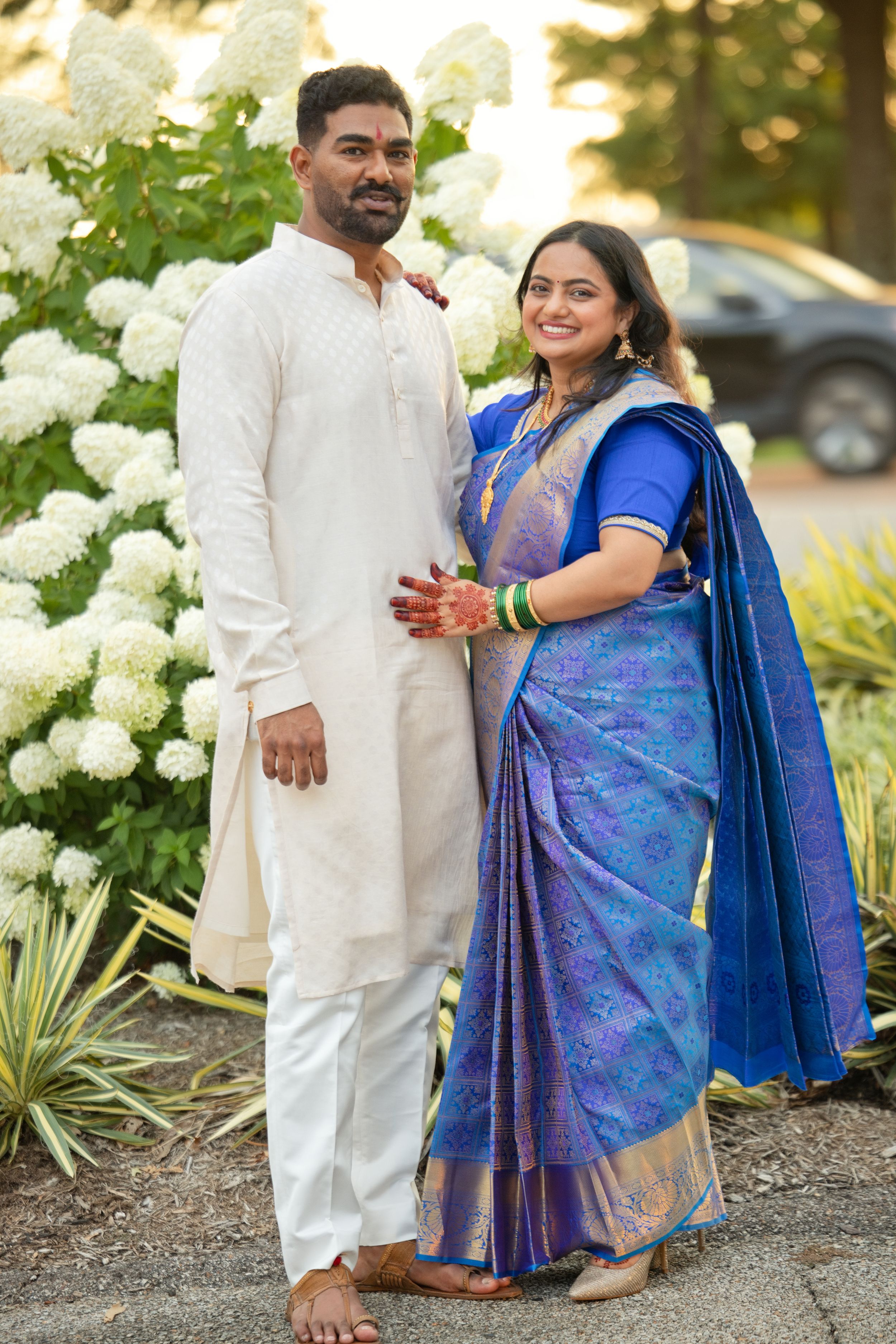Indian couple in traditional attire, man in white kurta and woman in blue silk saree, posing outdoors near white hydrangeas.