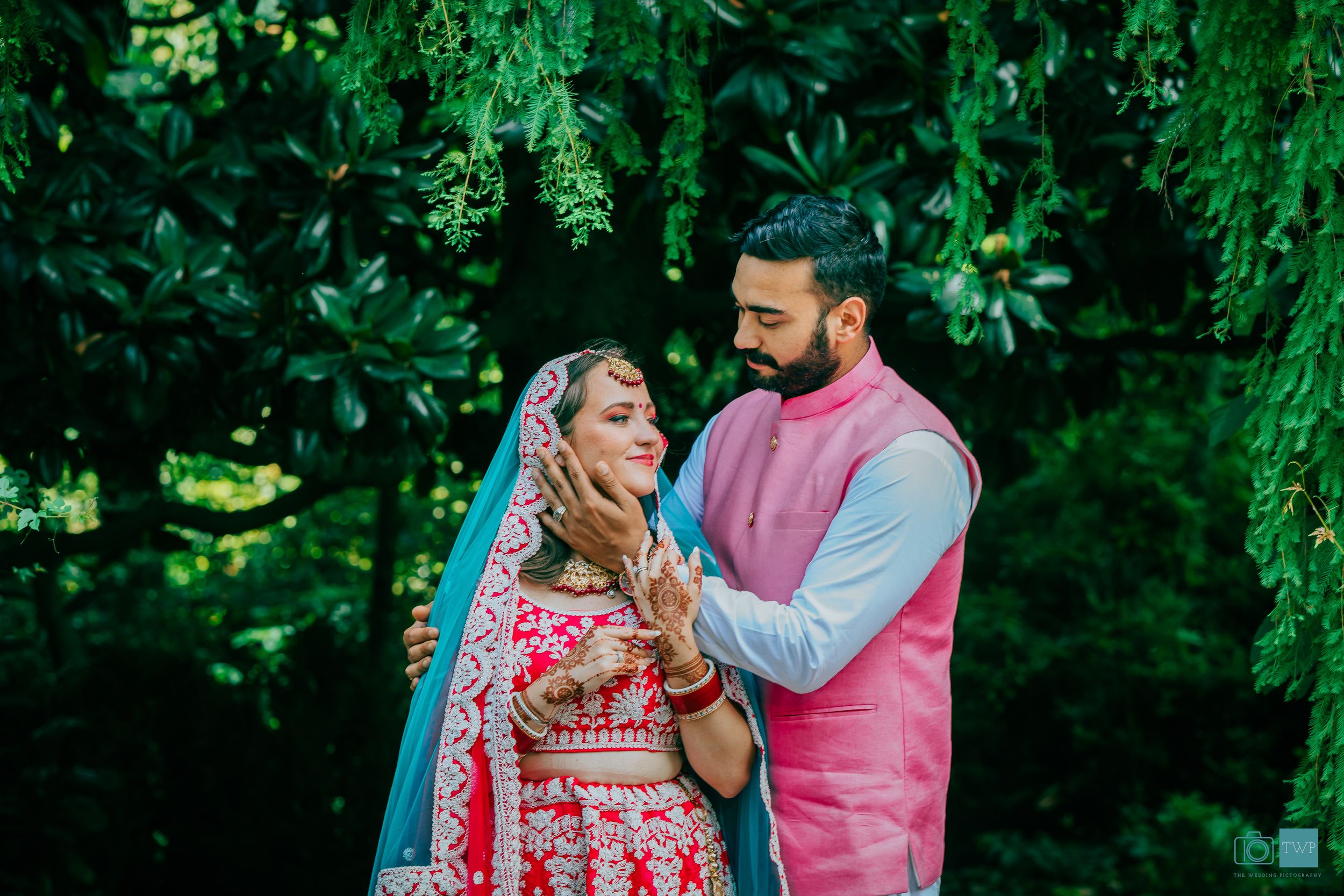 Indian bride and groom in traditional attire share a romantic moment surrounded by lush green foliage.