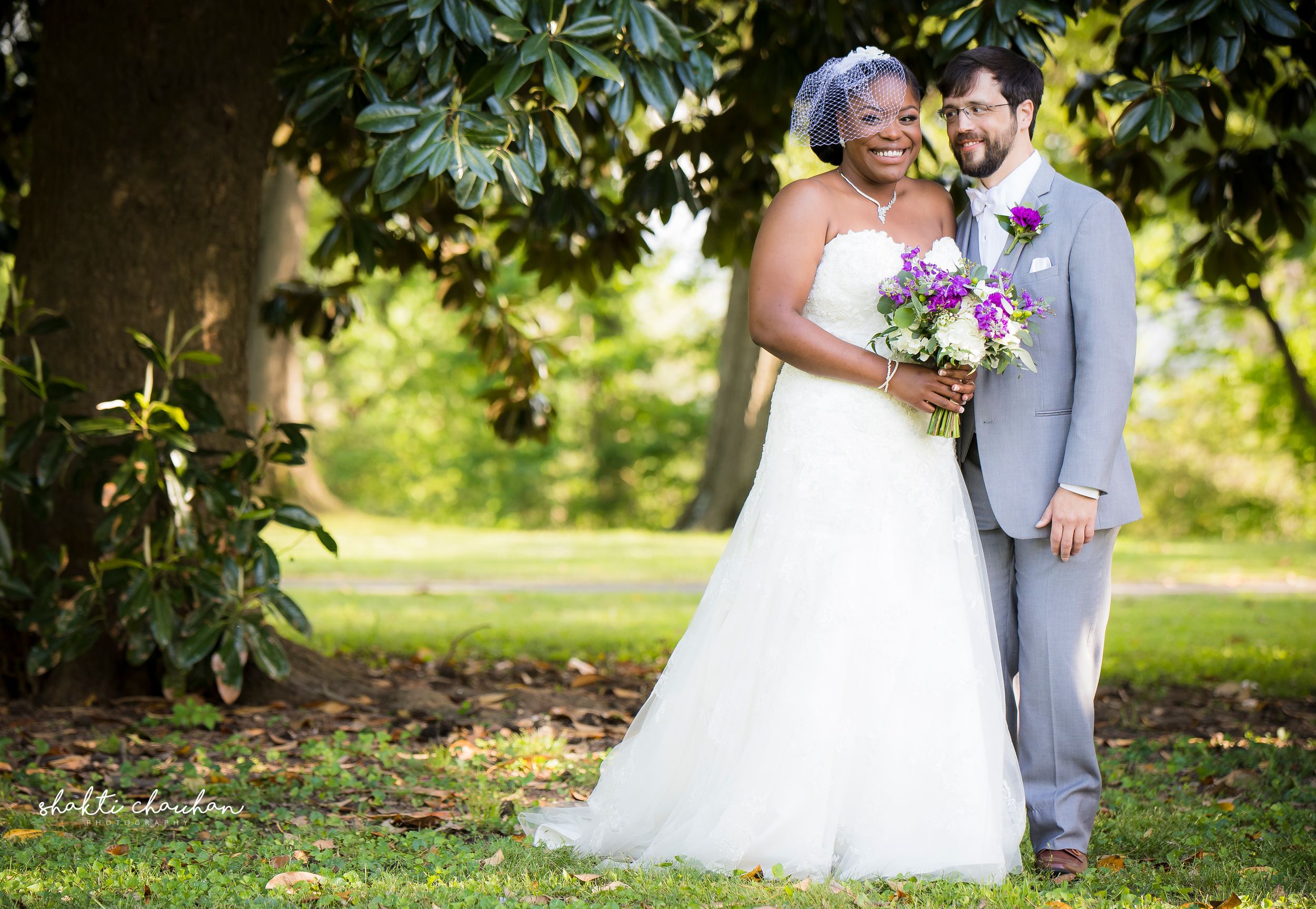 Interracial couple poses in lush garden on wedding day, bride in white gown with purple bouquet, groom in gray suit.