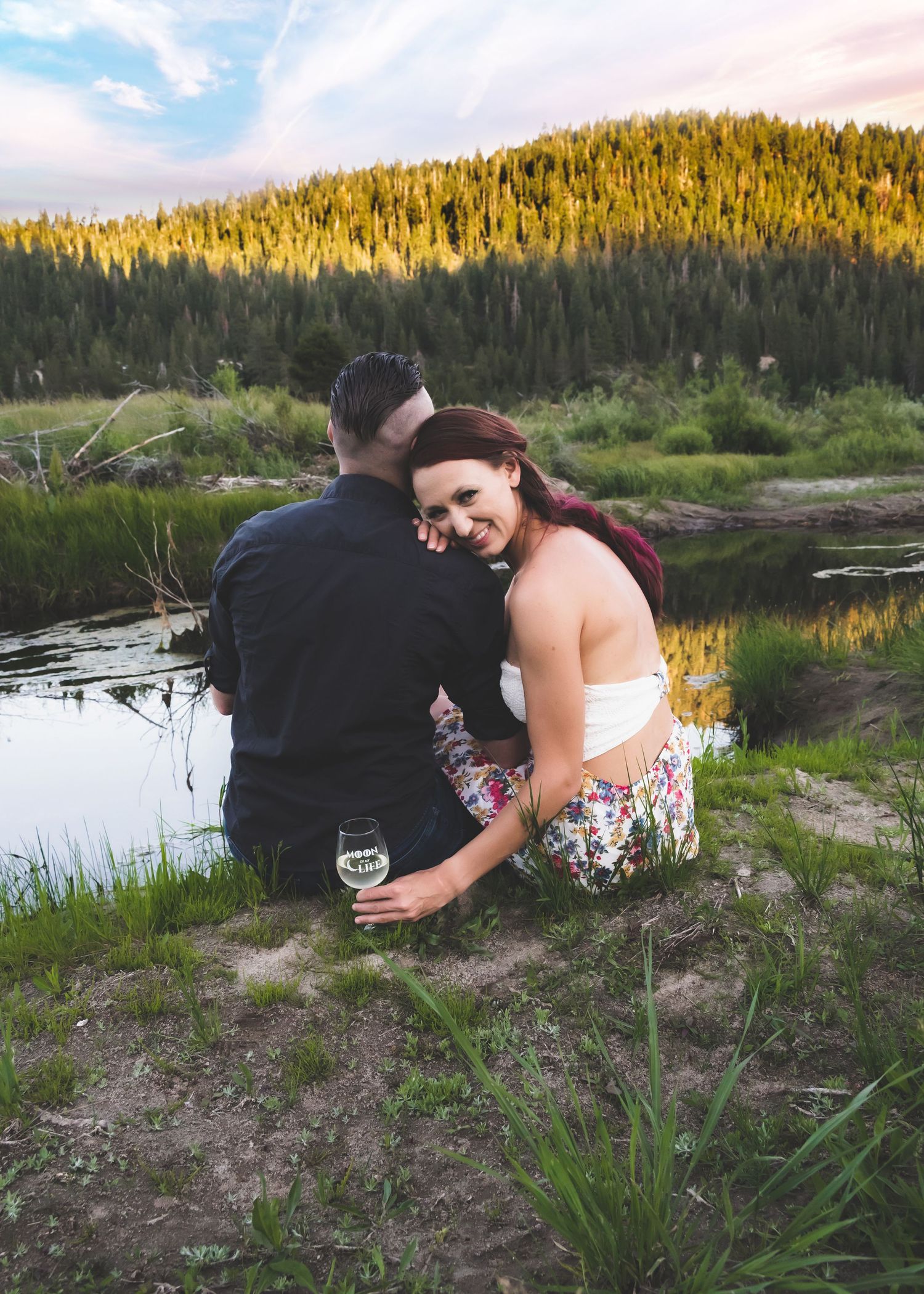 Couple sitting by a mountain lake at sunset, woman smiling holding wine glass, surrounded by pine trees and green grass.