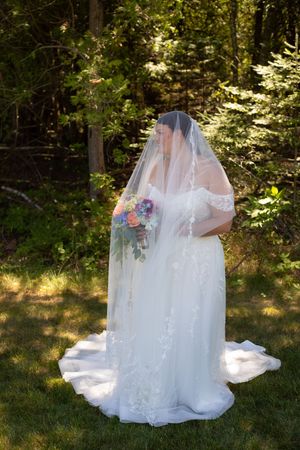 Bride in white lace gown and veil holding colorful bouquet standing outdoors in sunlit green forest setting.