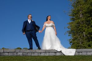 Bride in white gown and groom in navy suit pose on stone steps under a clear blue sky at outdoor wedding.