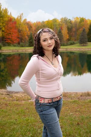 A person in a pink sweater and jeans poses playfully by a scenic autumn lake with colorful fall foliage in the background.