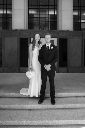 Black and white wedding photo showing couple in formal attire standing on stone steps outside a building.