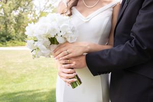 A couple in formal wedding attire holds a white floral bouquet during an outdoor ceremony.