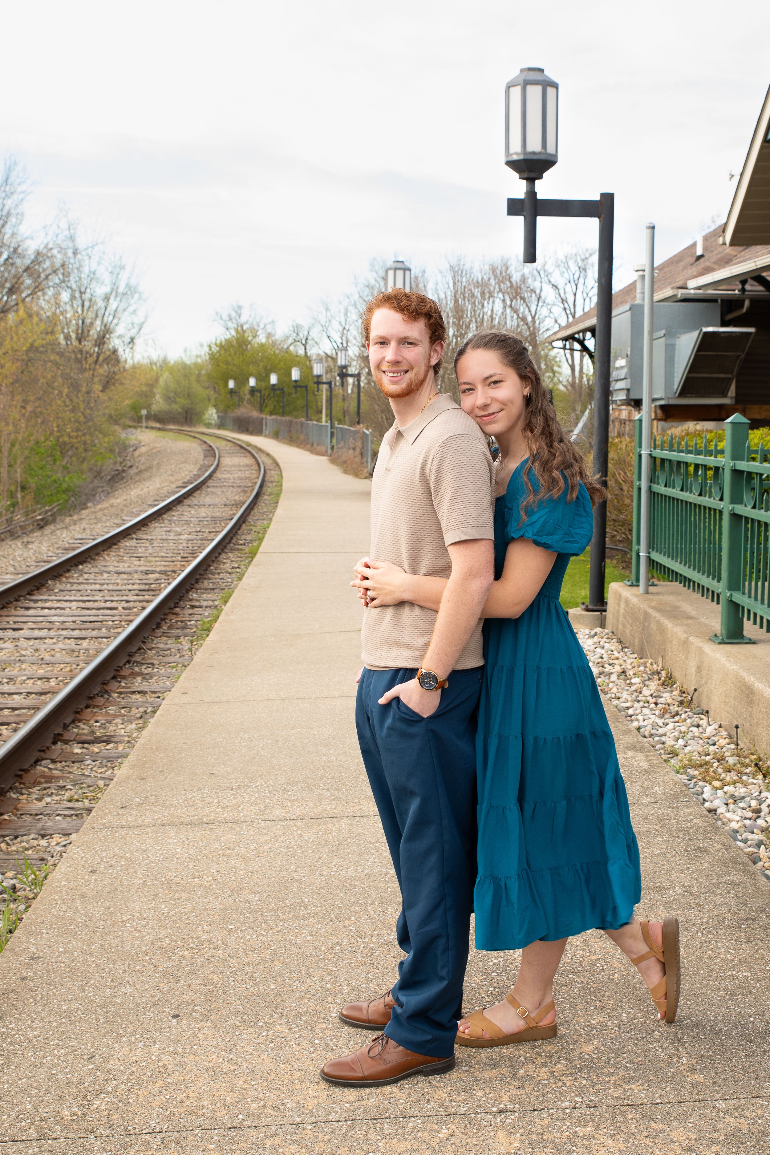A couple embraces near a curved train track and green railing during an outdoor engagement photoshoot.