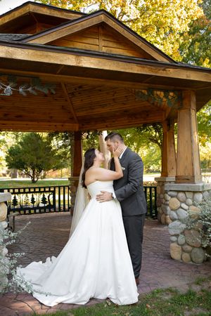 A romantic wedding couple shares intimate moments under a rustic wooden pavilion surrounded by autumn foliage.