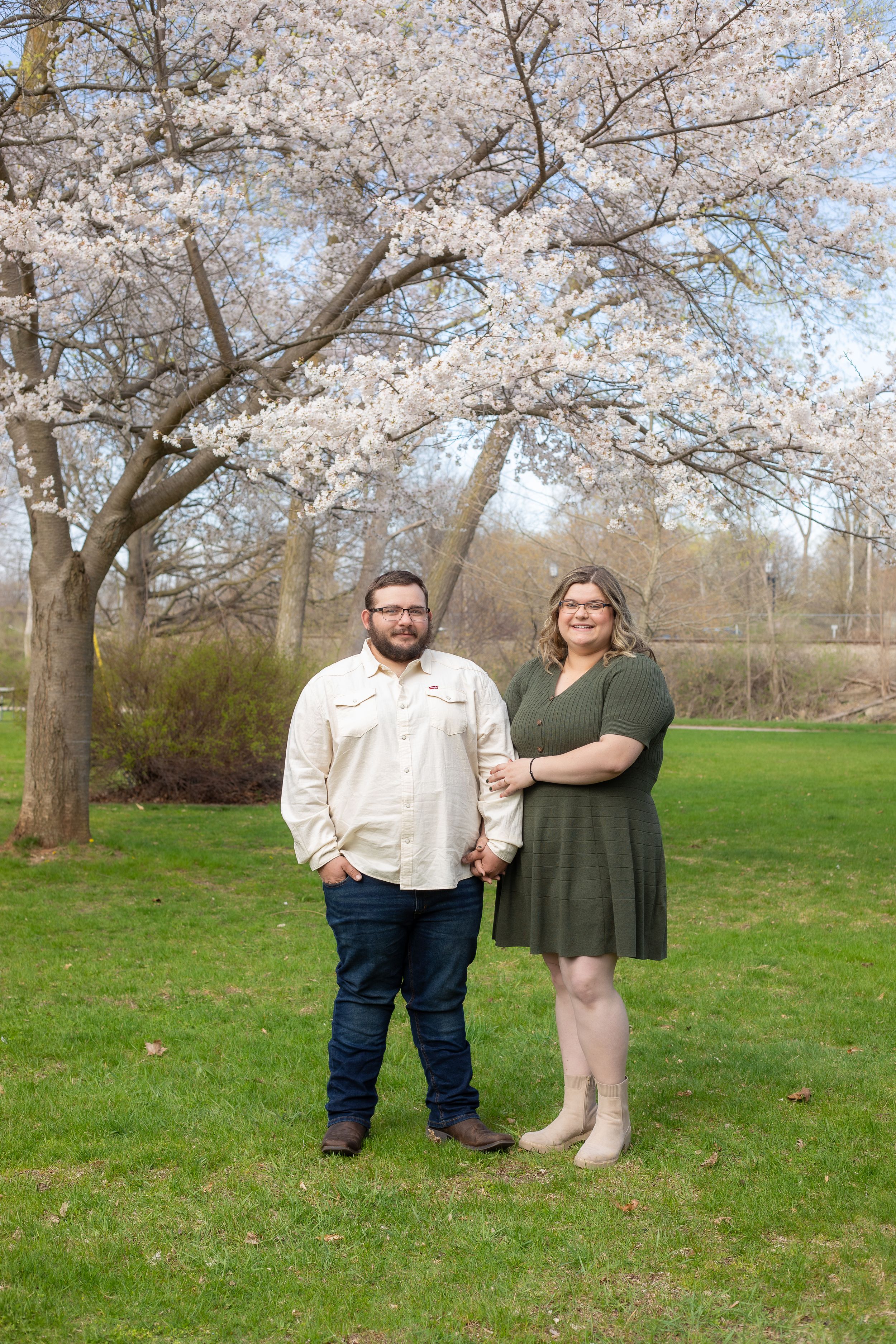 A couple poses for a portrait beneath flowering cherry blossom trees on a grassy lawn.
