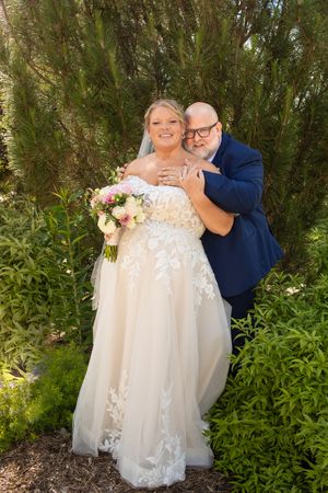A bride in lace gown holding pink bouquet embraced by groom in navy suit among lush green garden foliage.