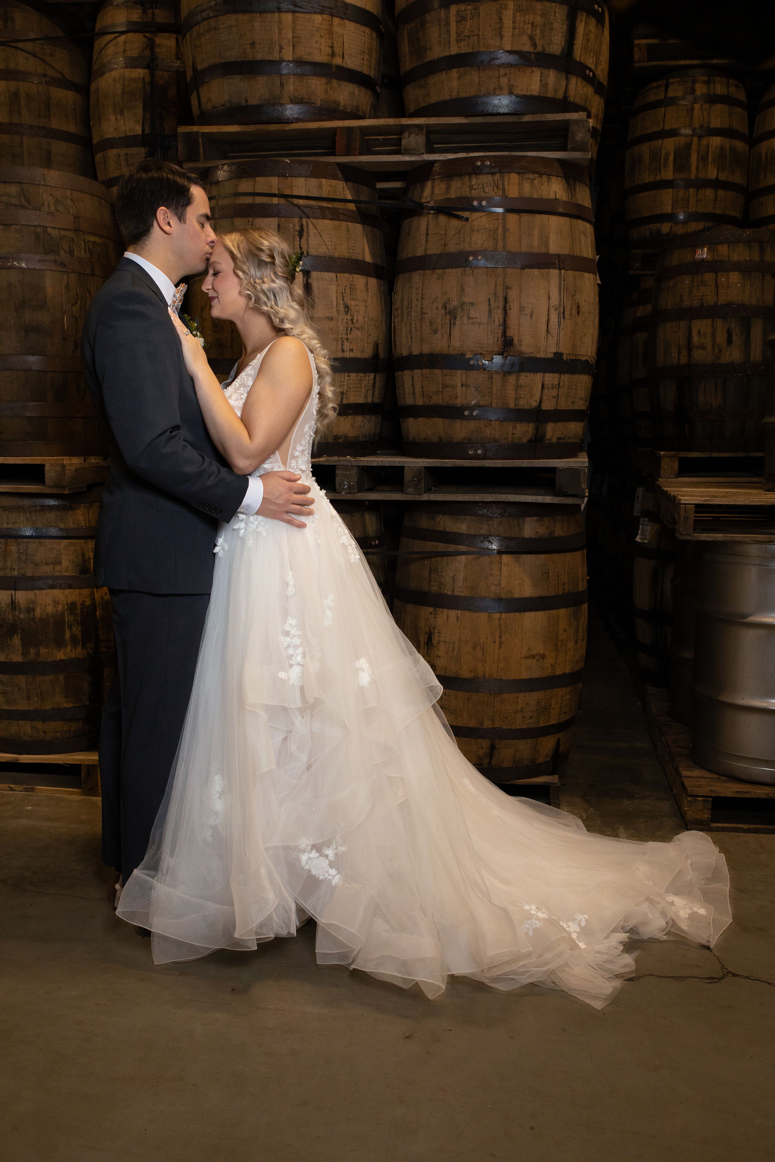 A romantic wedding photo featuring stacked wooden barrels in the background and a flowing white dress.