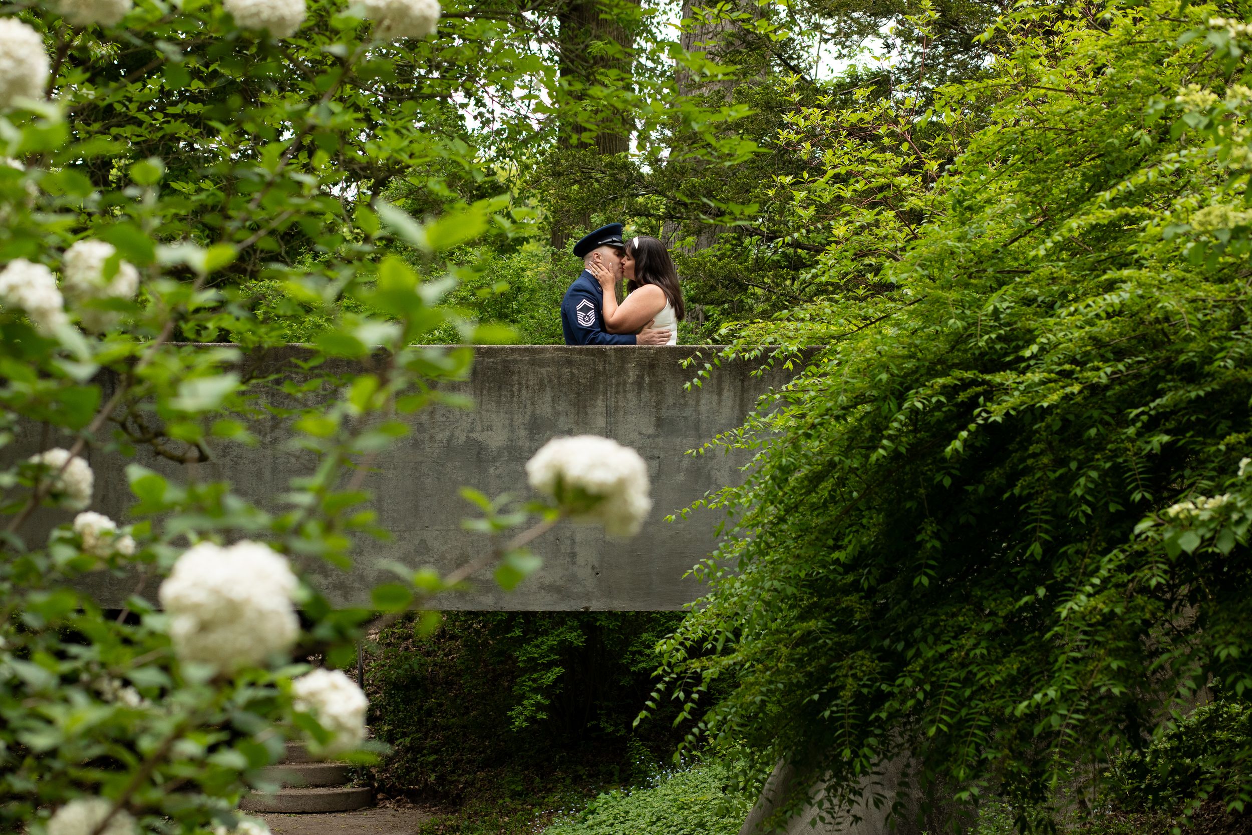 A couple embraces on a romantic wooden footbridge surrounded by lush green foliage and white blooming flowers.