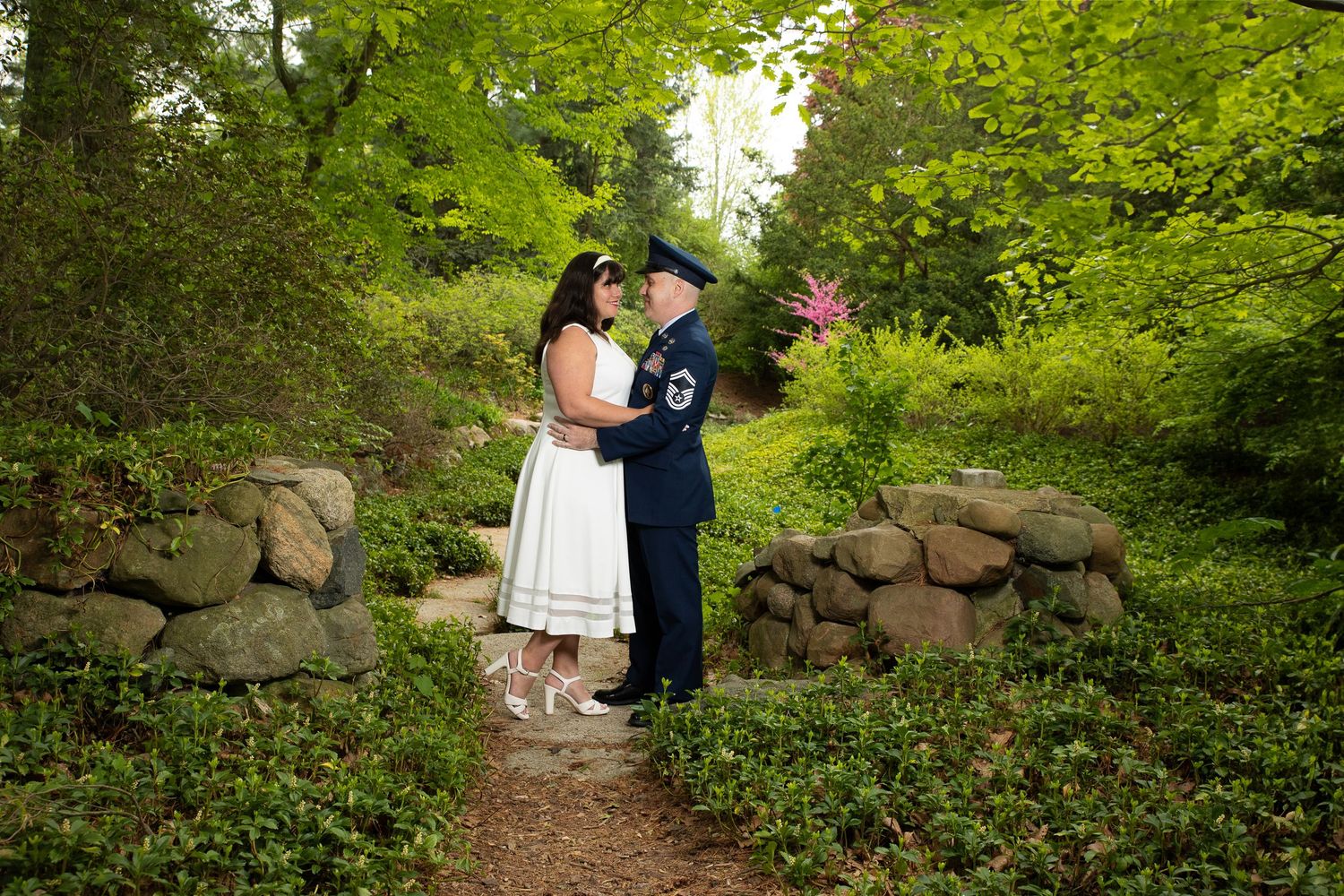 Two people share an intimate moment on a garden path surrounded by lush greenery and stone walls.
