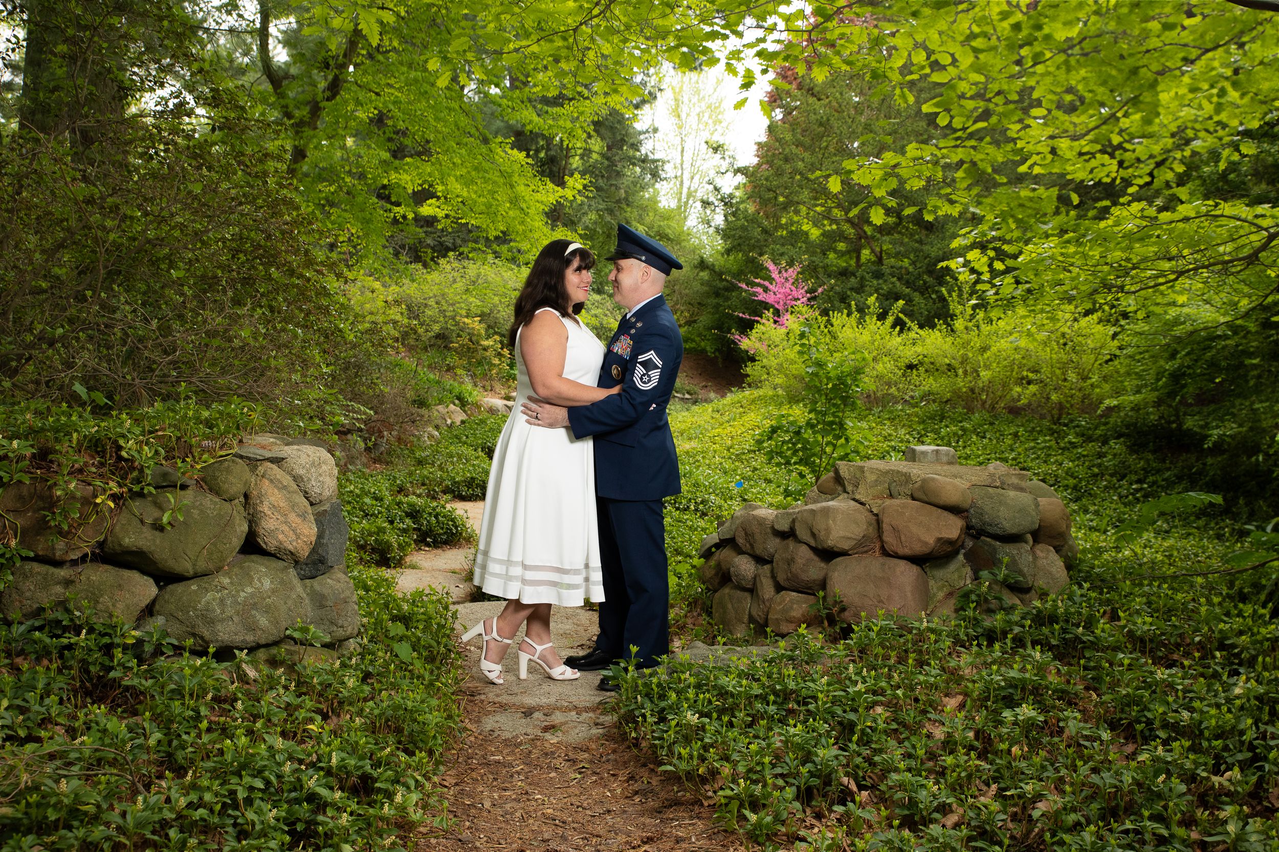 Two people share an intimate moment on a garden path surrounded by lush greenery and stone walls.