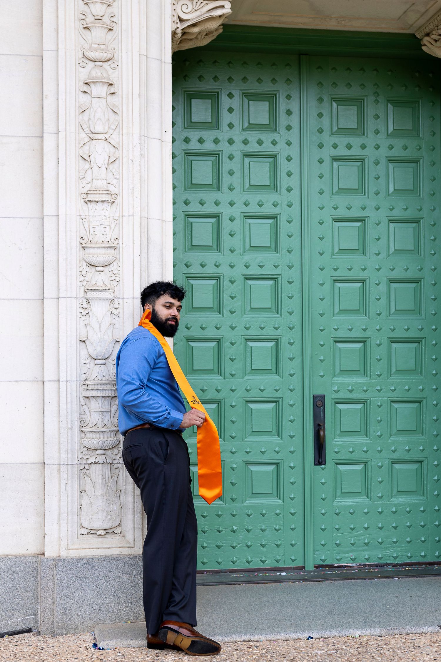 Person in blue jacket and orange accessory stands against large ornate mint green door with decorative white stone columns.