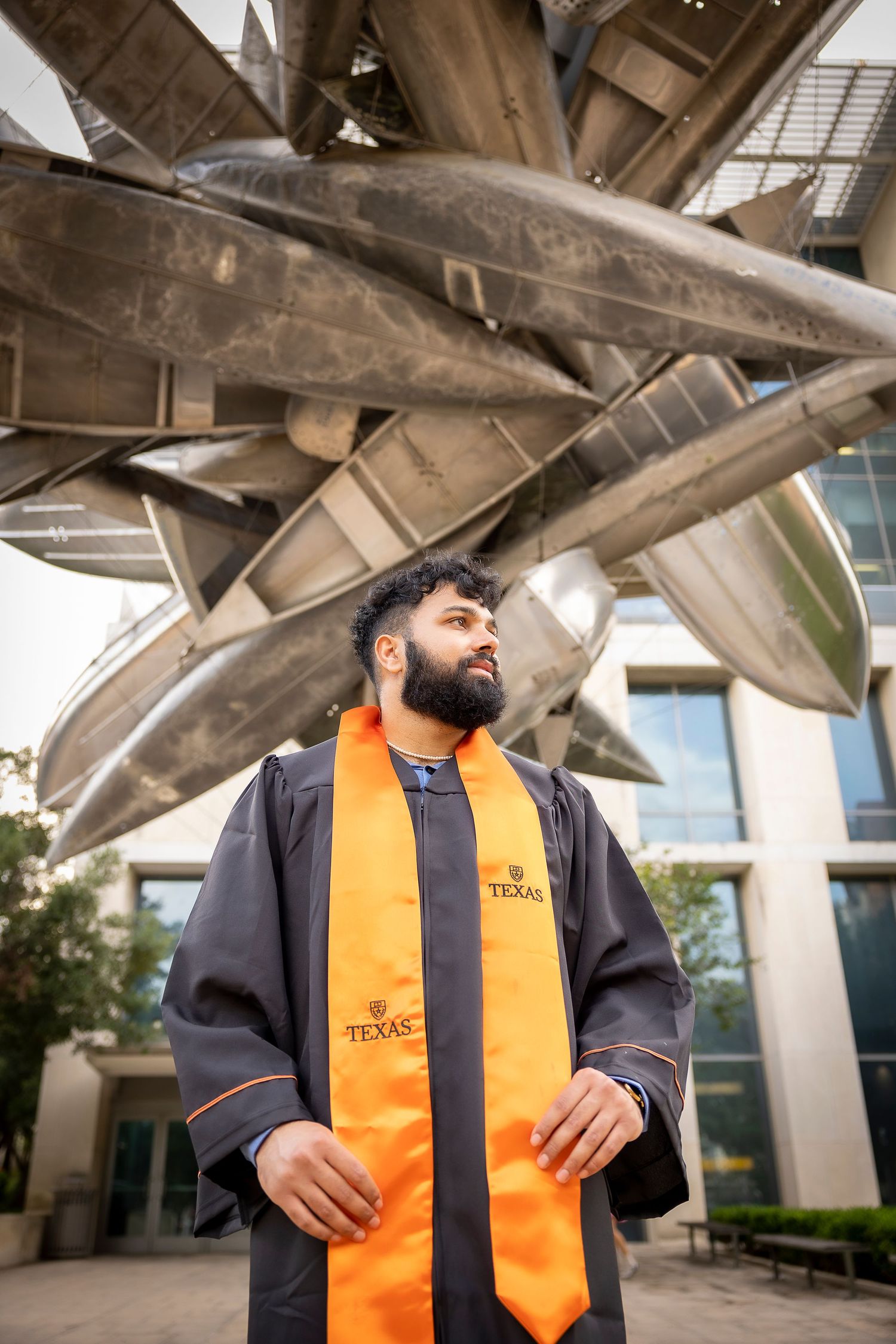 A graduate in orange academic stole and navy gown stands under modern architectural beams.