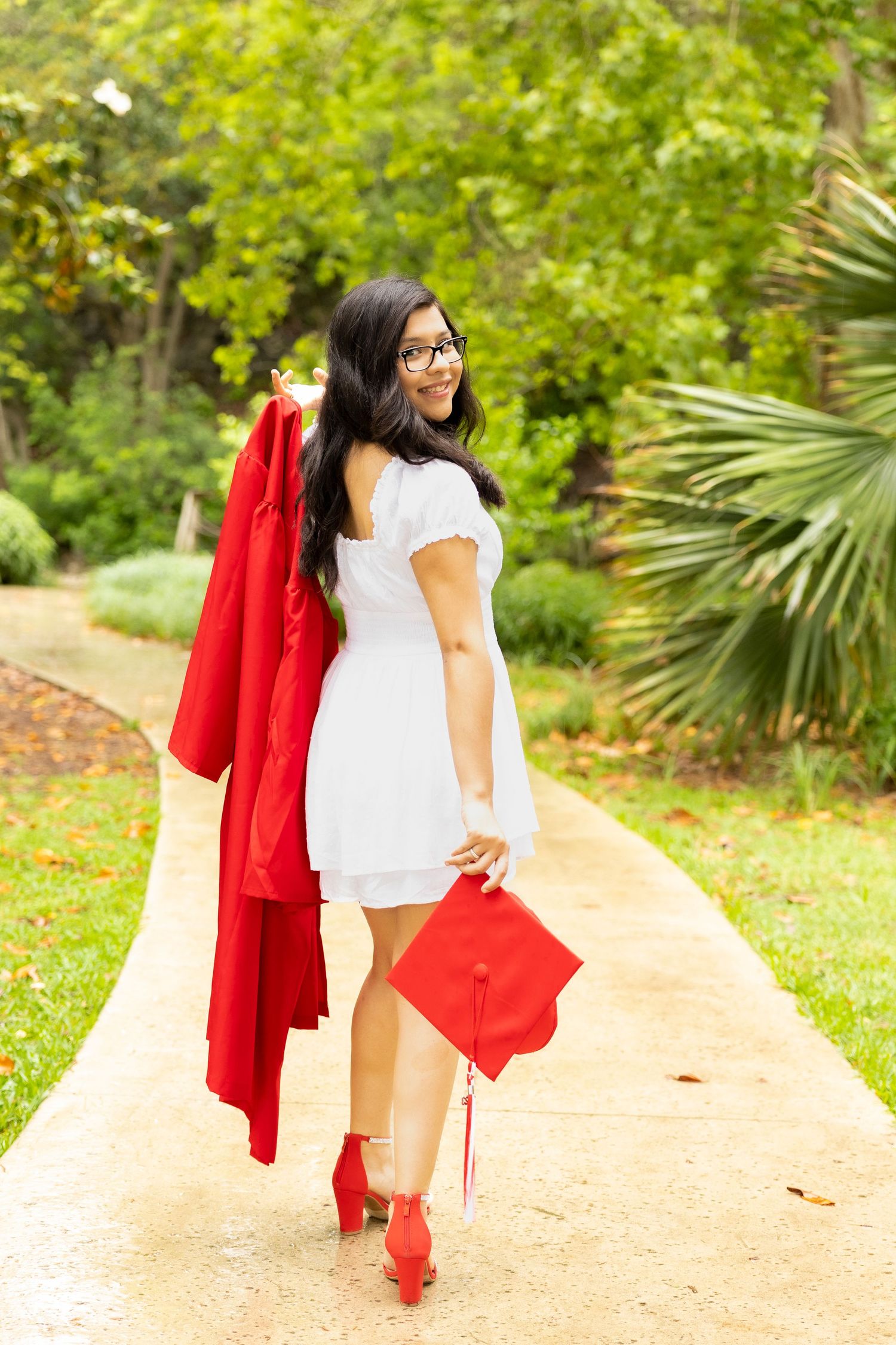 Graduate in white dress and red graduation gown holds cap while walking on outdoor path surrounded by greenery.