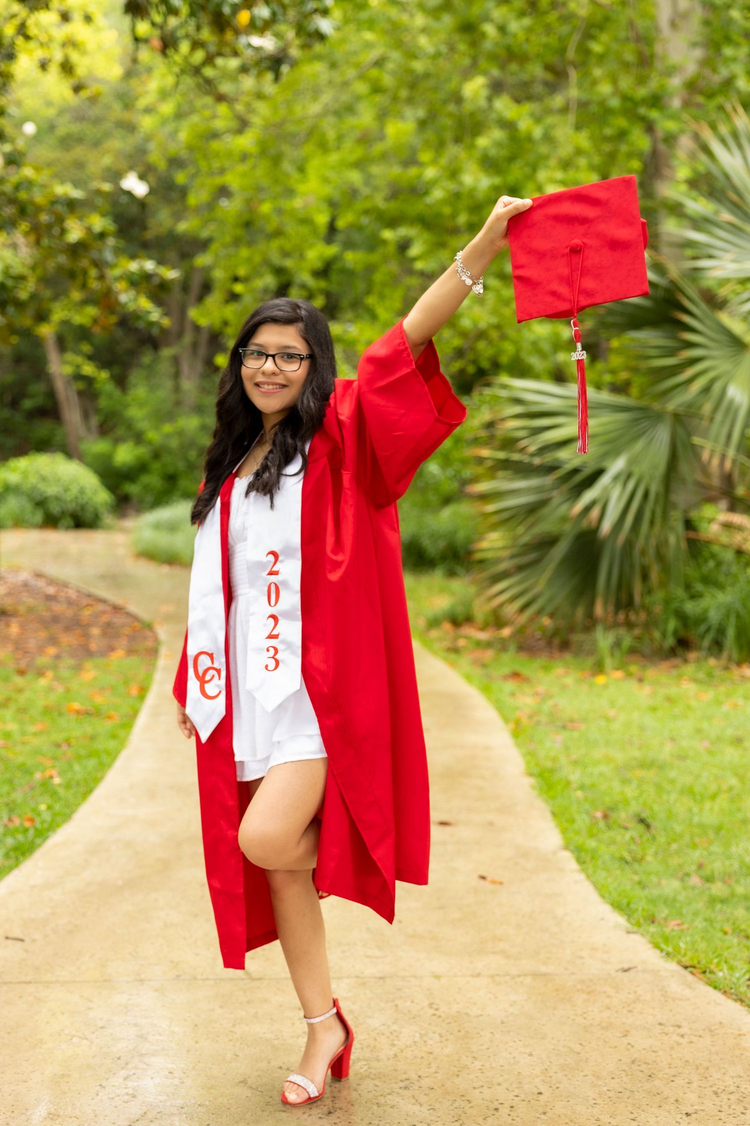 A graduate in red cap and gown poses for celebratory photos on a dirt path surrounded by lush greenery.