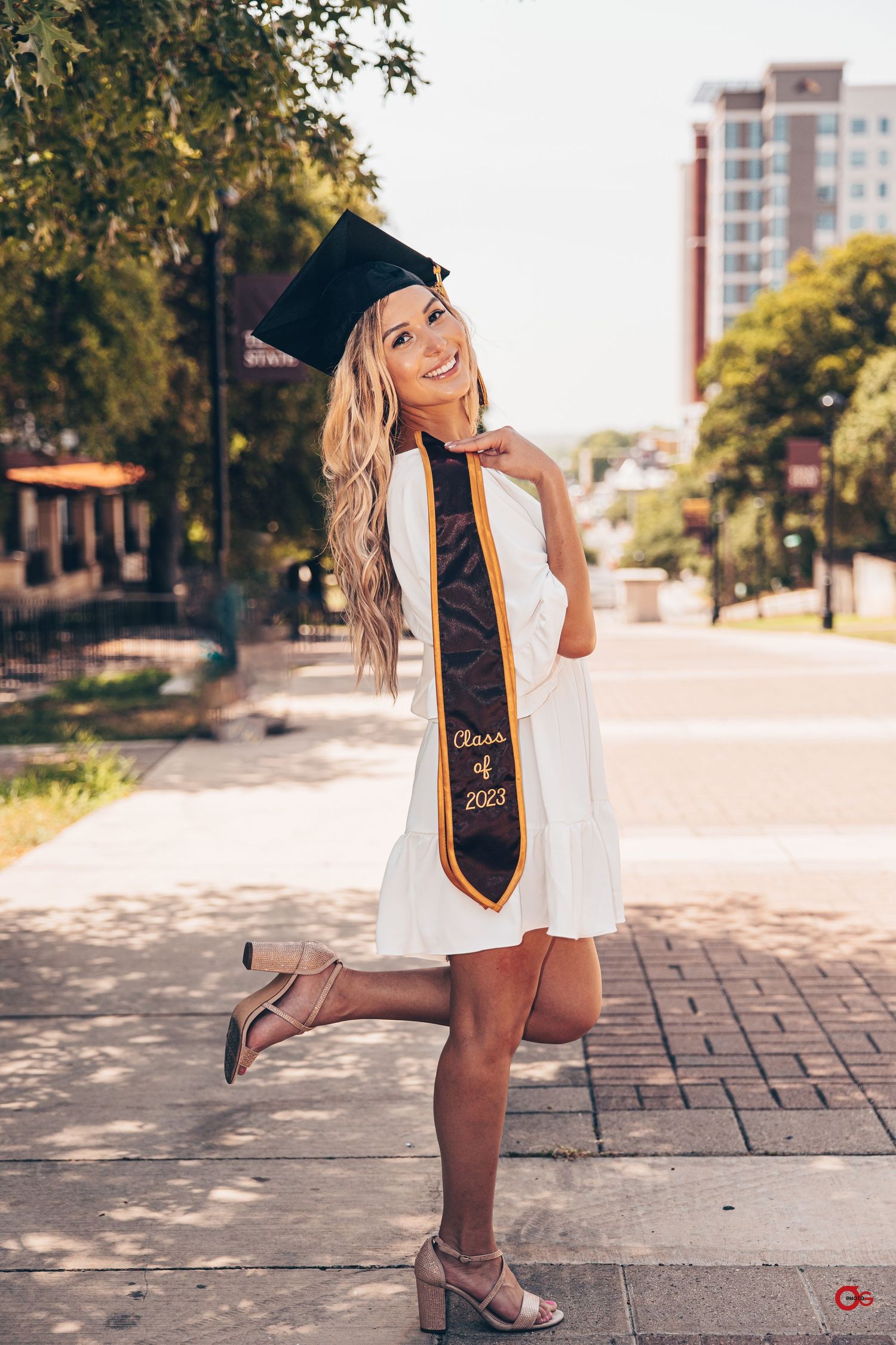 Graduate in white dress and cap poses on brick pathway with graduation sash during sunny outdoor photoshoot.