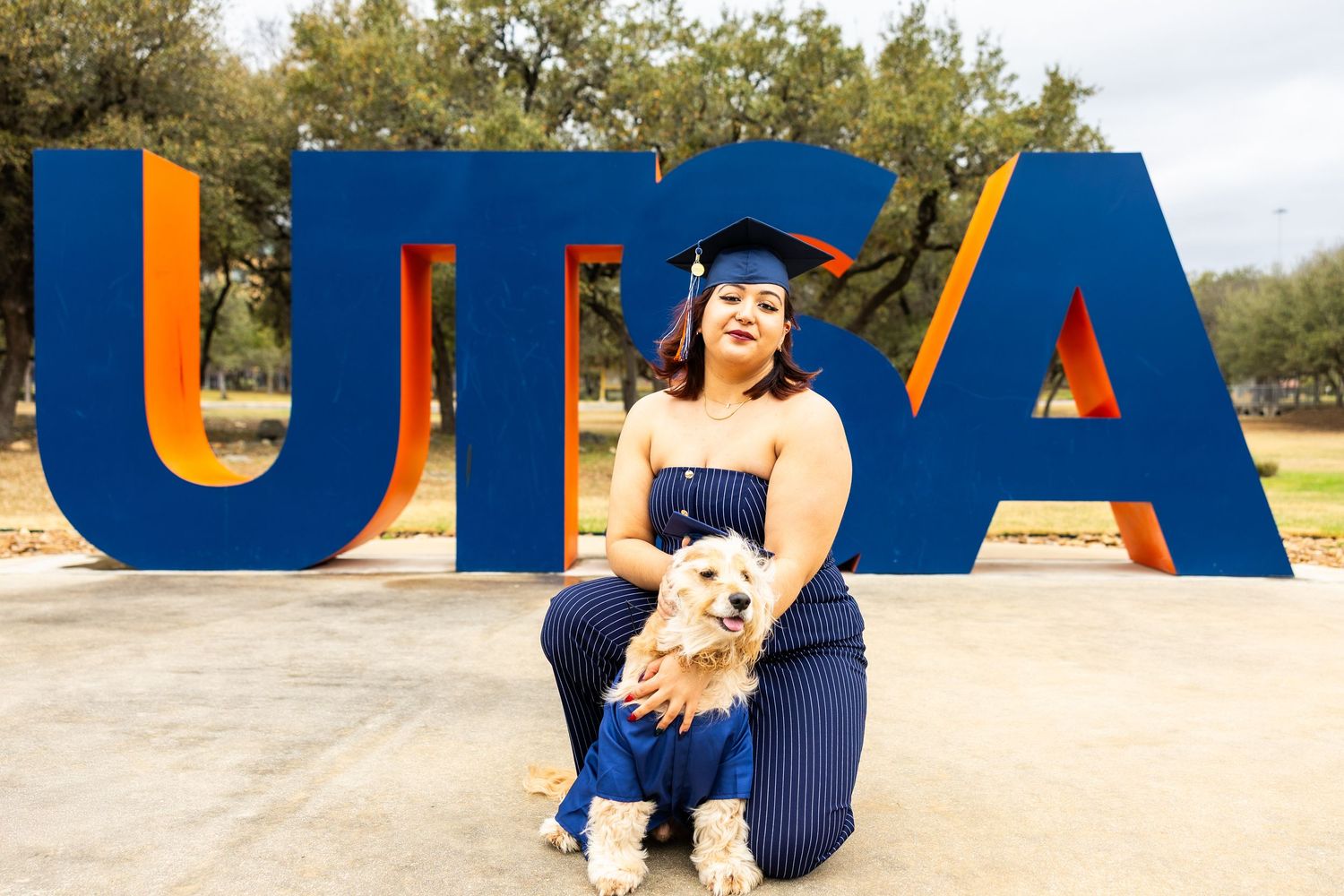 A person stands next to large blue letters spelling 'UTSA' outdoors on a sunny day.