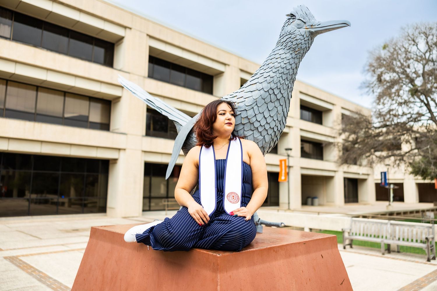 A joyful figure in blue sits on a brick ledge near a modern building with a large decorative bird sculpture.