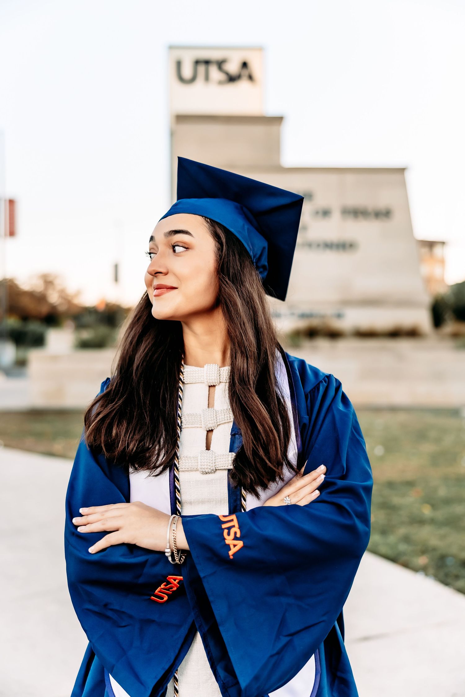Graduate in blue cap and gown poses outdoors on campus with UTSA building visible in background.