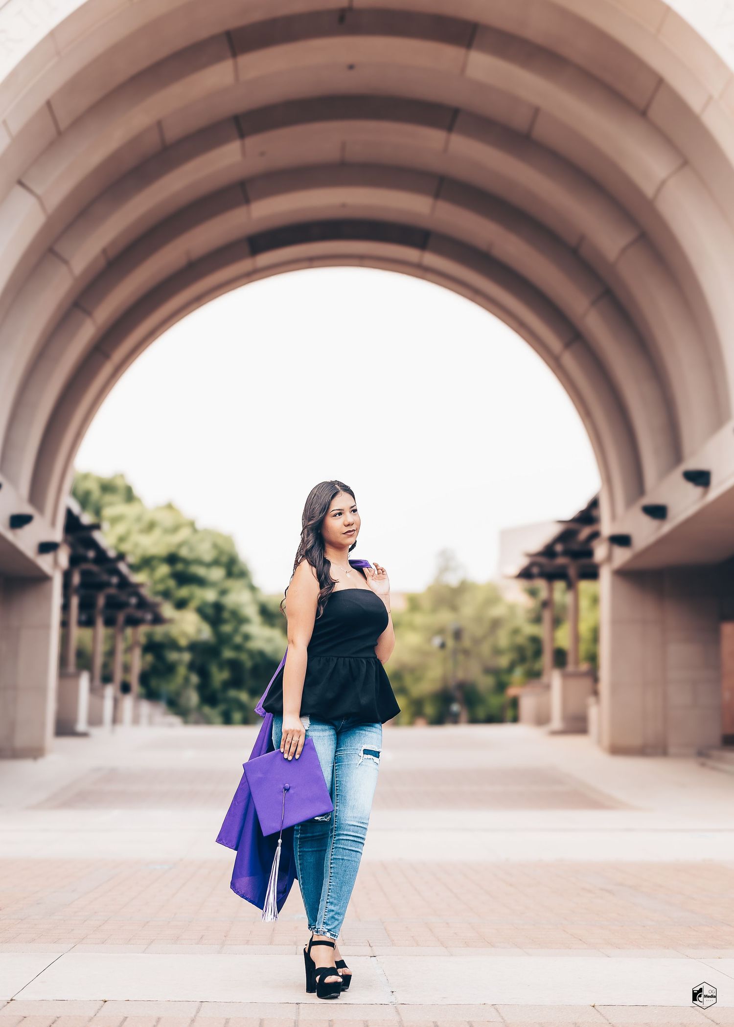 Fashionable woman in black peplum top and jeans poses under concrete archway holding purple shopping bags.
