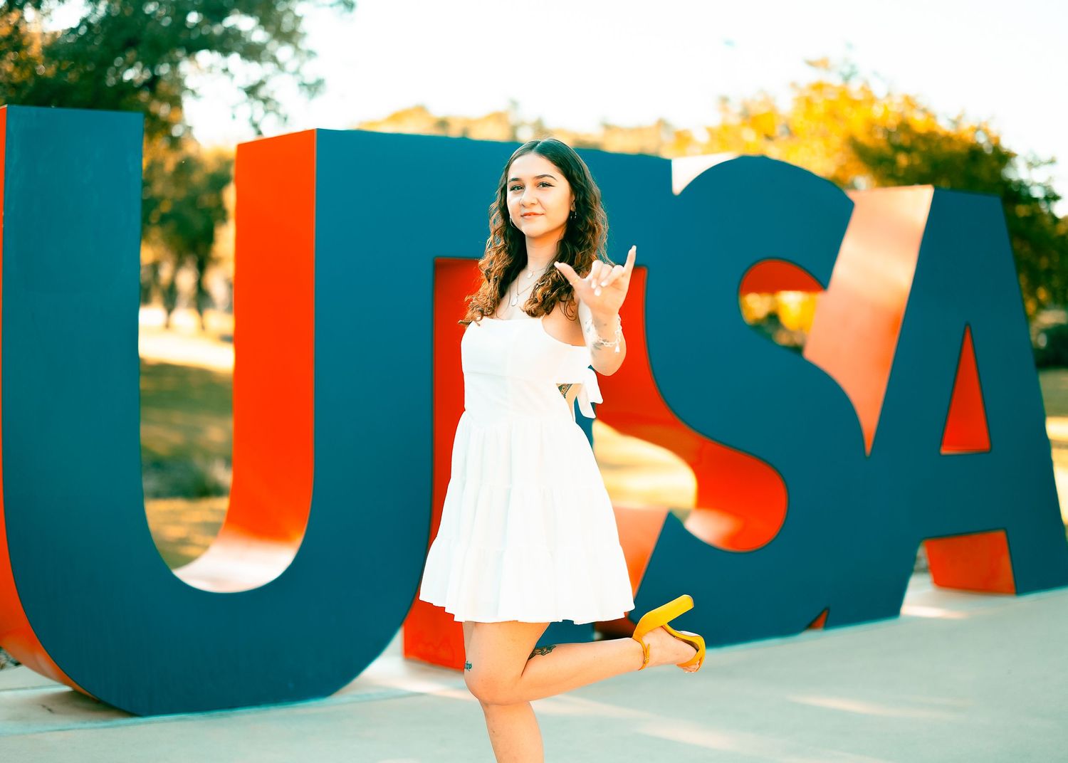 Someone in a white dress poses playfully in front of large red and blue USA letters outdoors during golden hour.