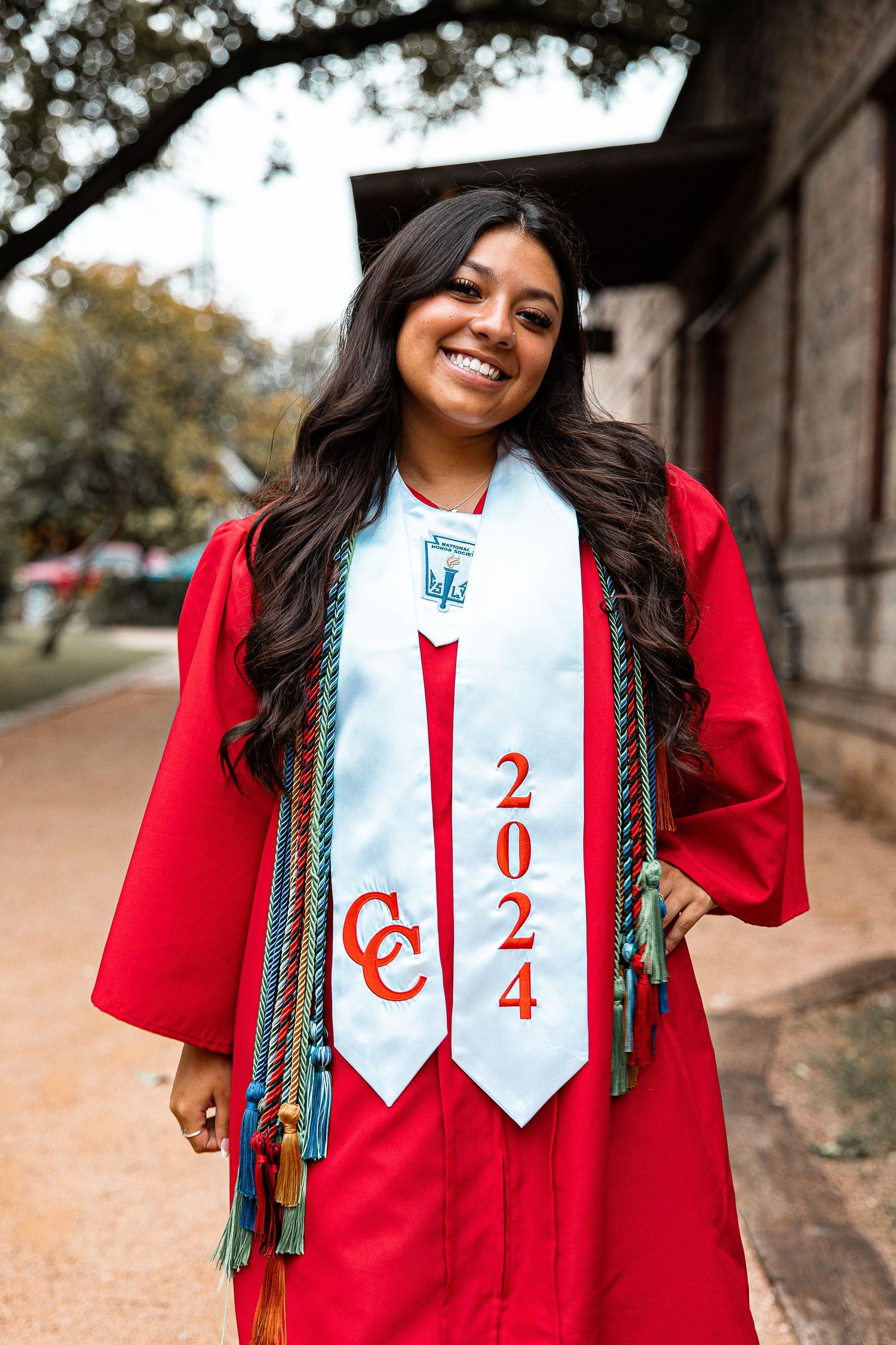 Graduate in red cap and gown wearing graduation stoles with decorative braids poses outdoors during commencement.