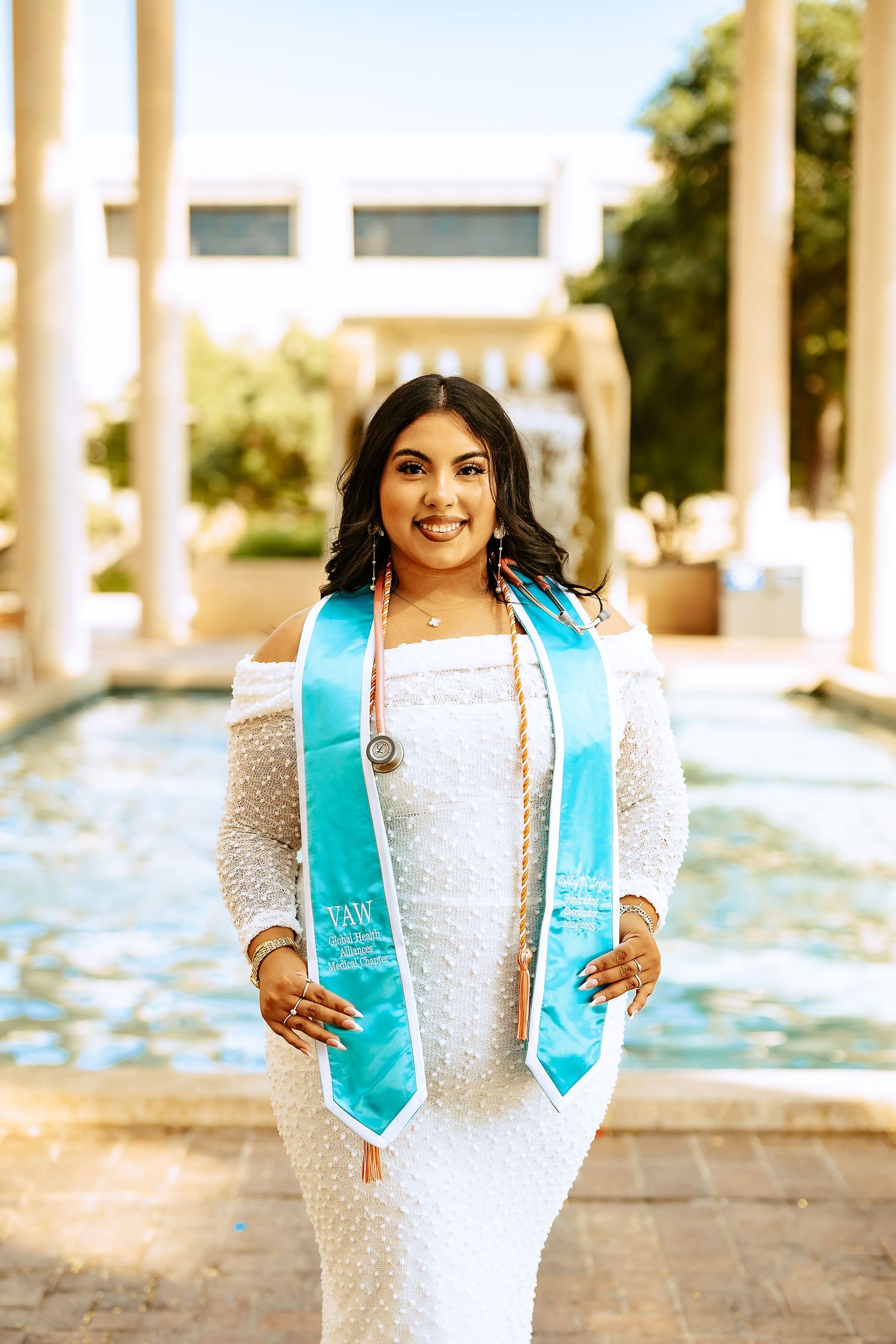 Graduate wearing white lace dress and turquoise graduation stole poses by fountain during outdoor ceremony.