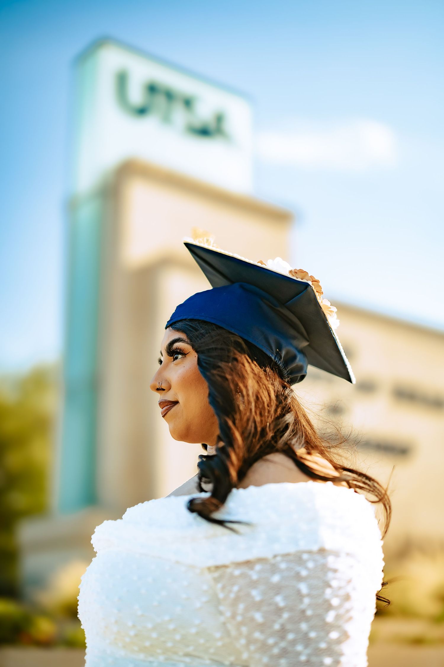 A graduate in a blue cap and white dress stands in profile against a blurred outdoor background.