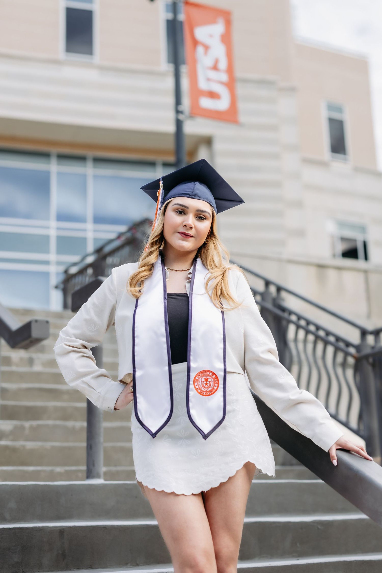 Graduate in white cap and gown poses on concrete steps near orange university banners.