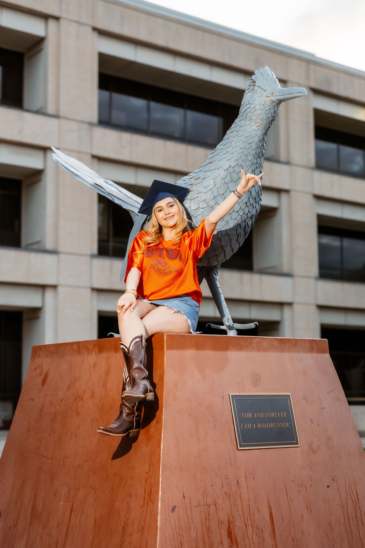 Graduate in orange shirt and graduation cap poses playfully on outdoor campus sculpture in multiple creative positions.