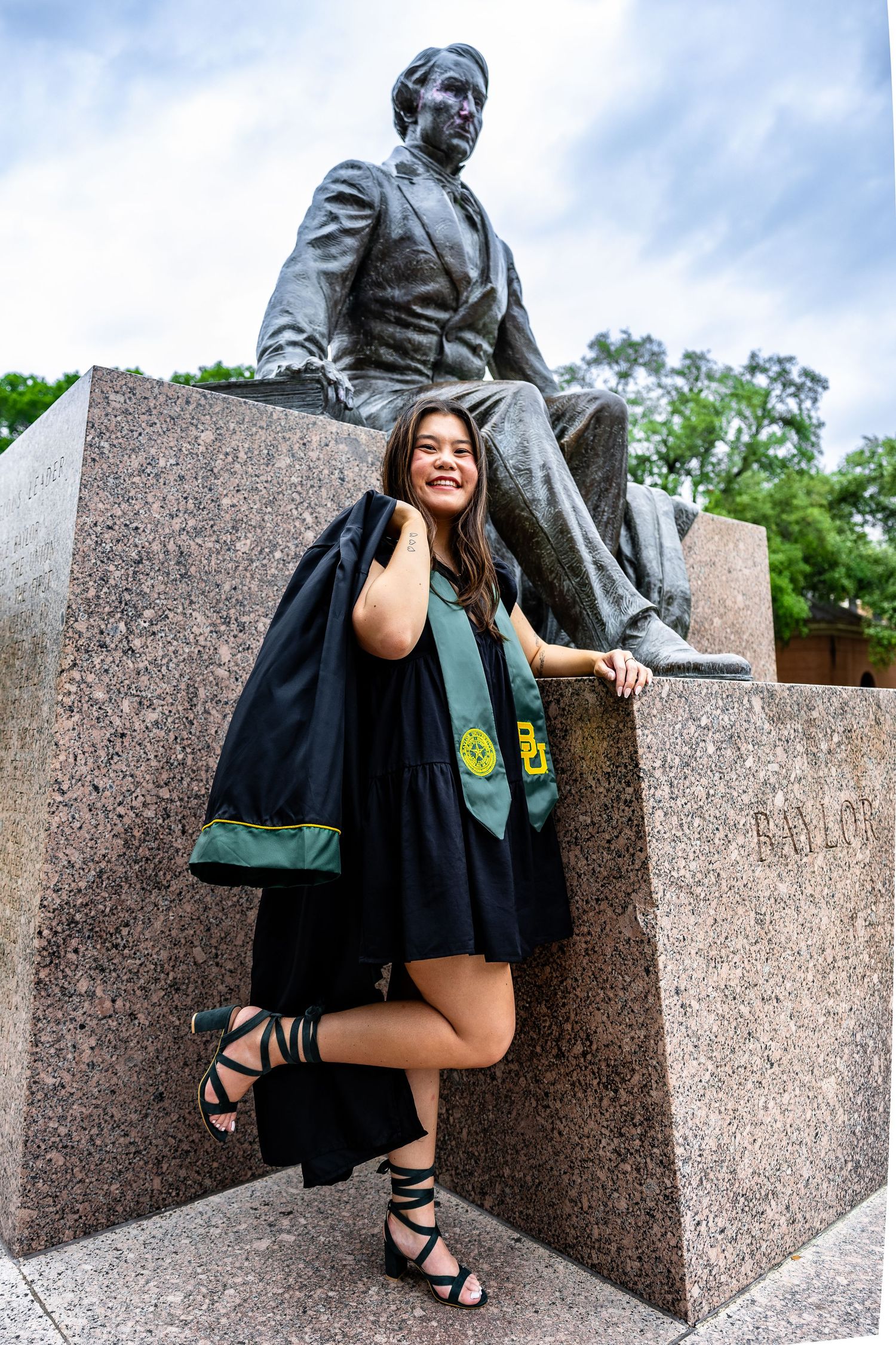 A graduate in black regalia poses with a bronze statue on a granite monument during a graduation photoshoot.