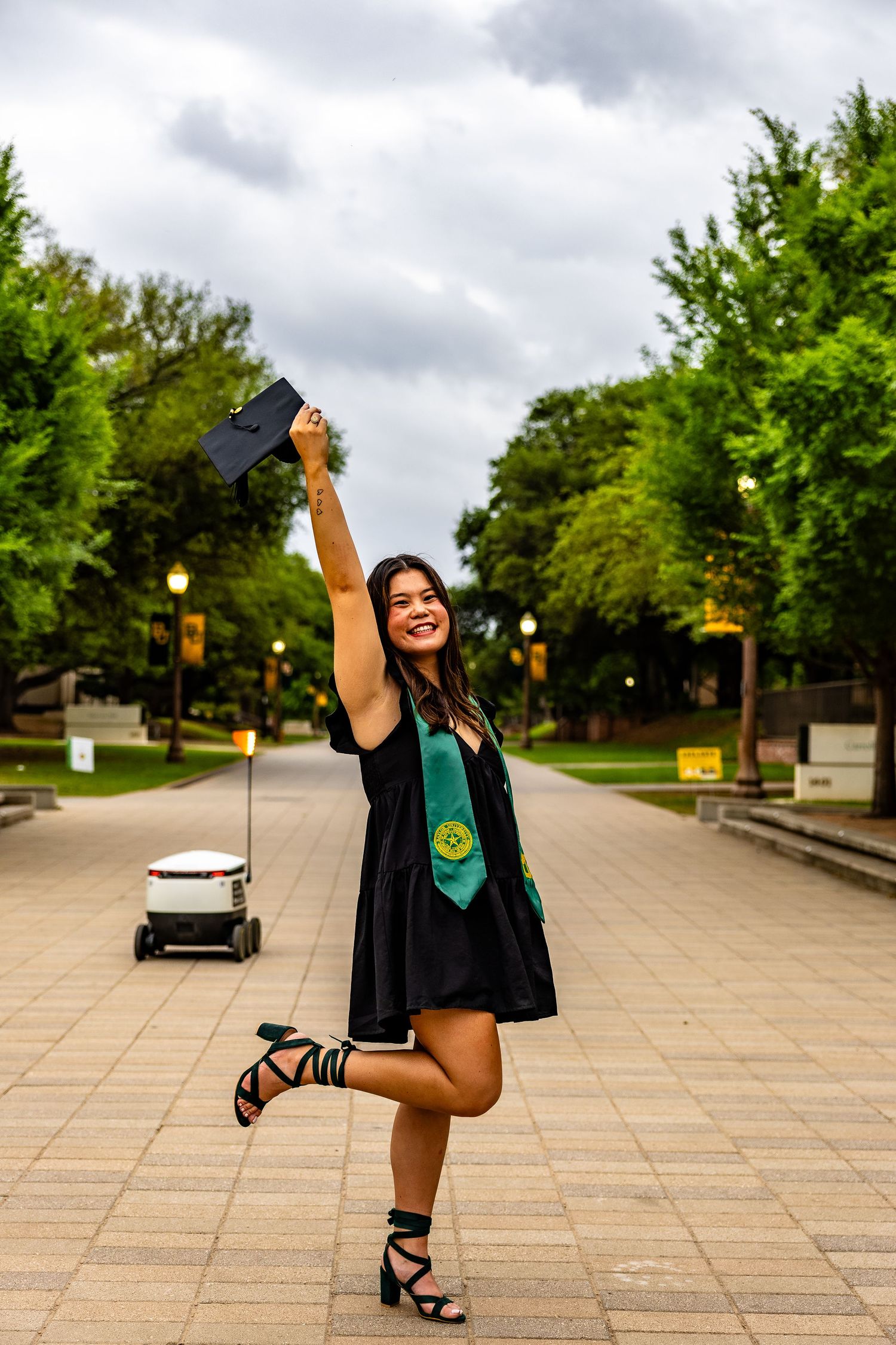 Graduate in cap and gown celebrates on campus walkway while striking joyful pose with graduation cap raised high.