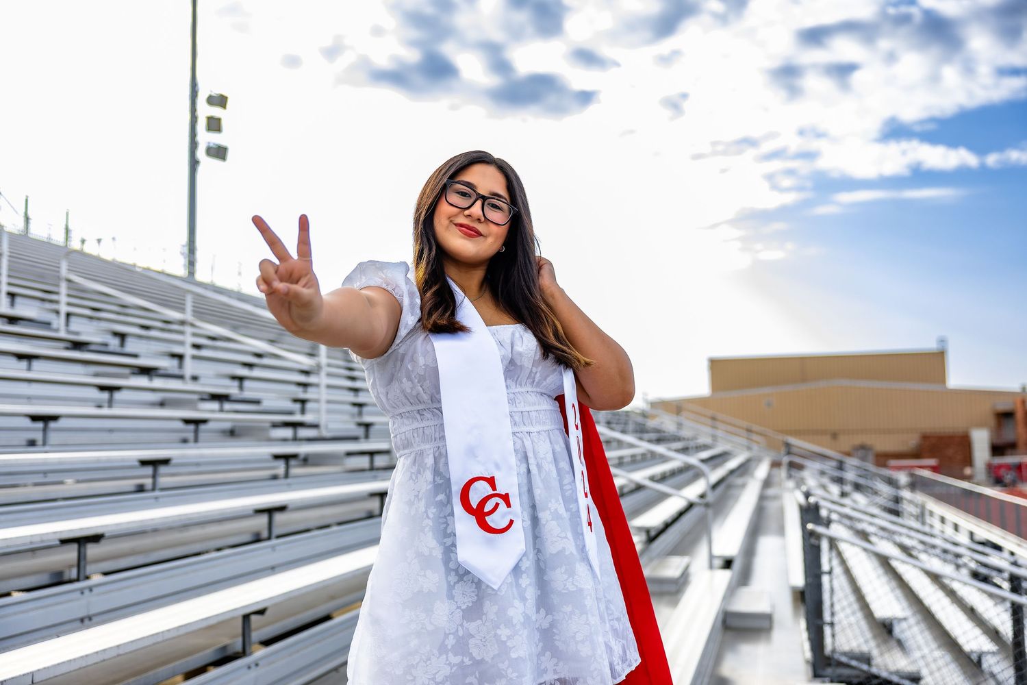 A person wearing a red and white outfit poses cheerfully on metal bleachers against a blue cloudy sky.