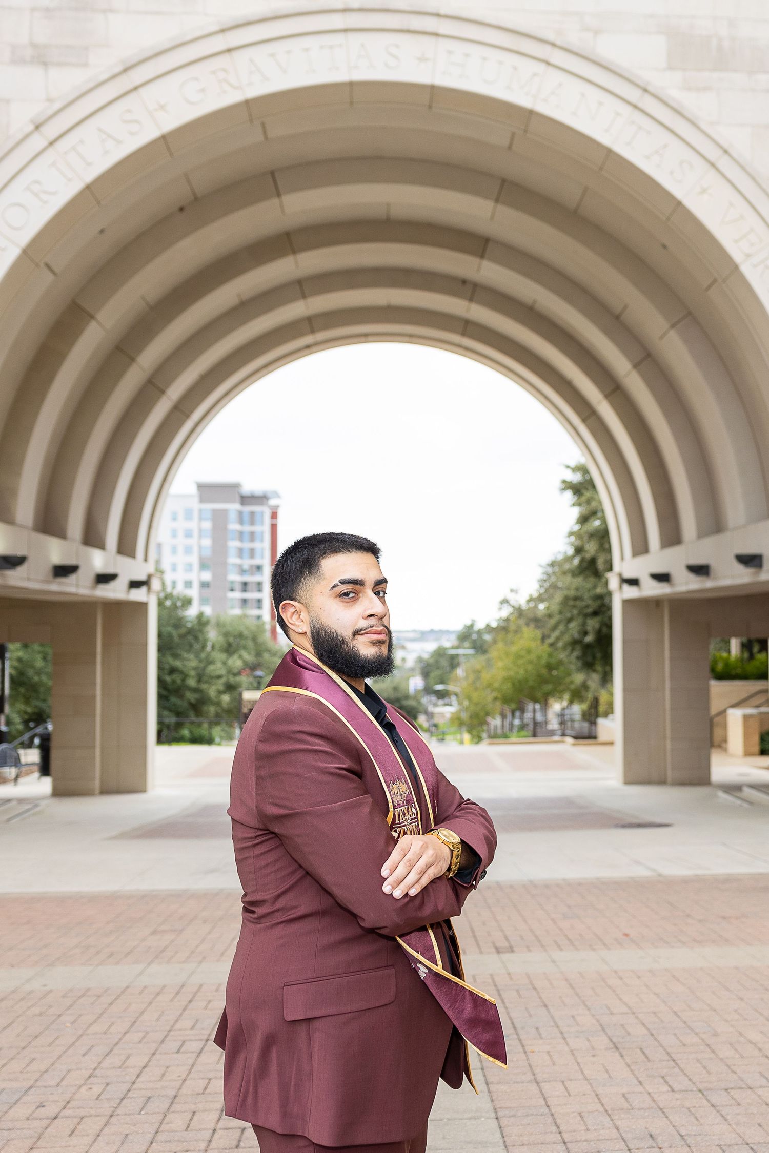 A person in a maroon suit poses beneath concrete arches in an architectural setting.