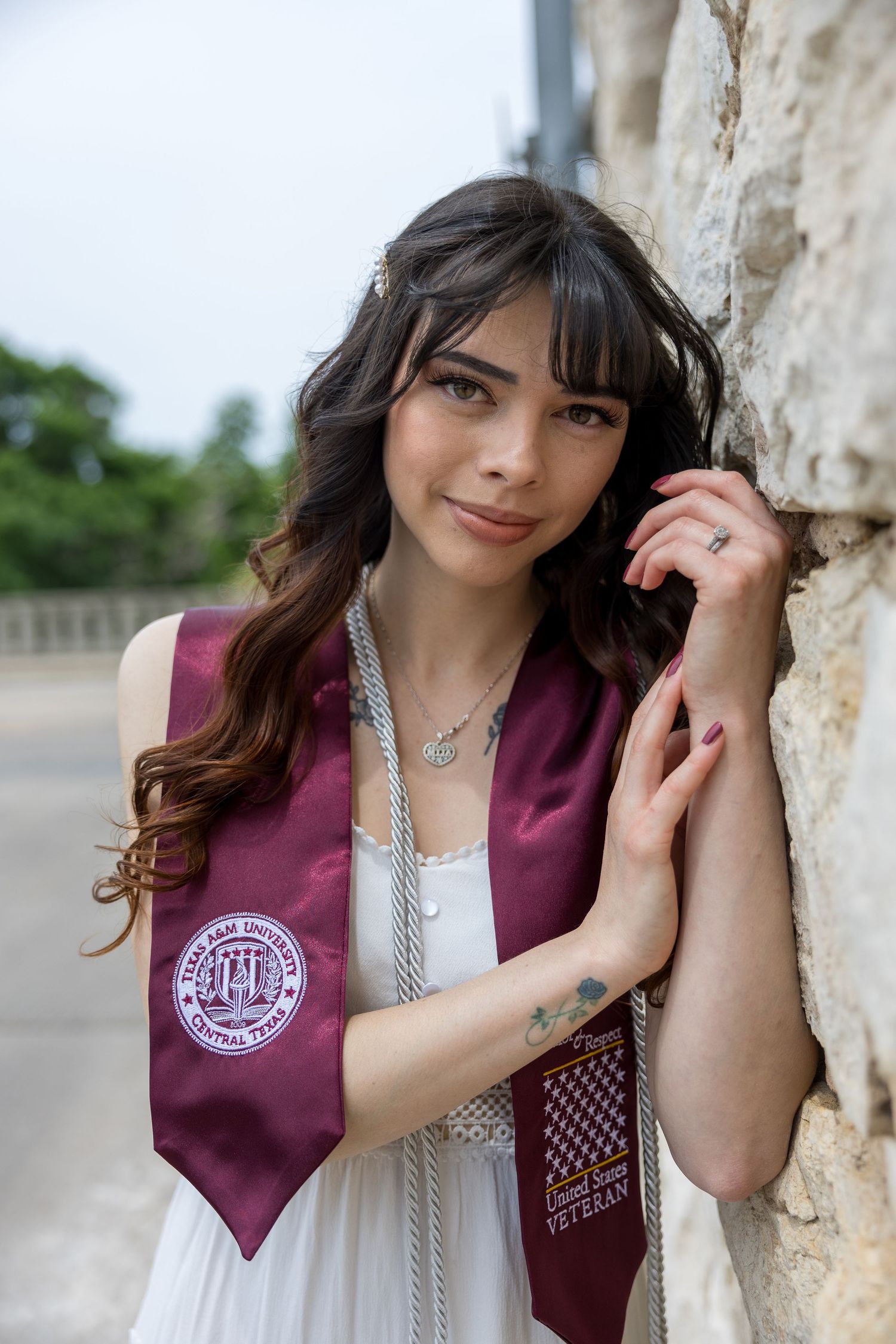 Graduate posing in maroon graduation stole with white lace dress against stone wall for senior portraits.
