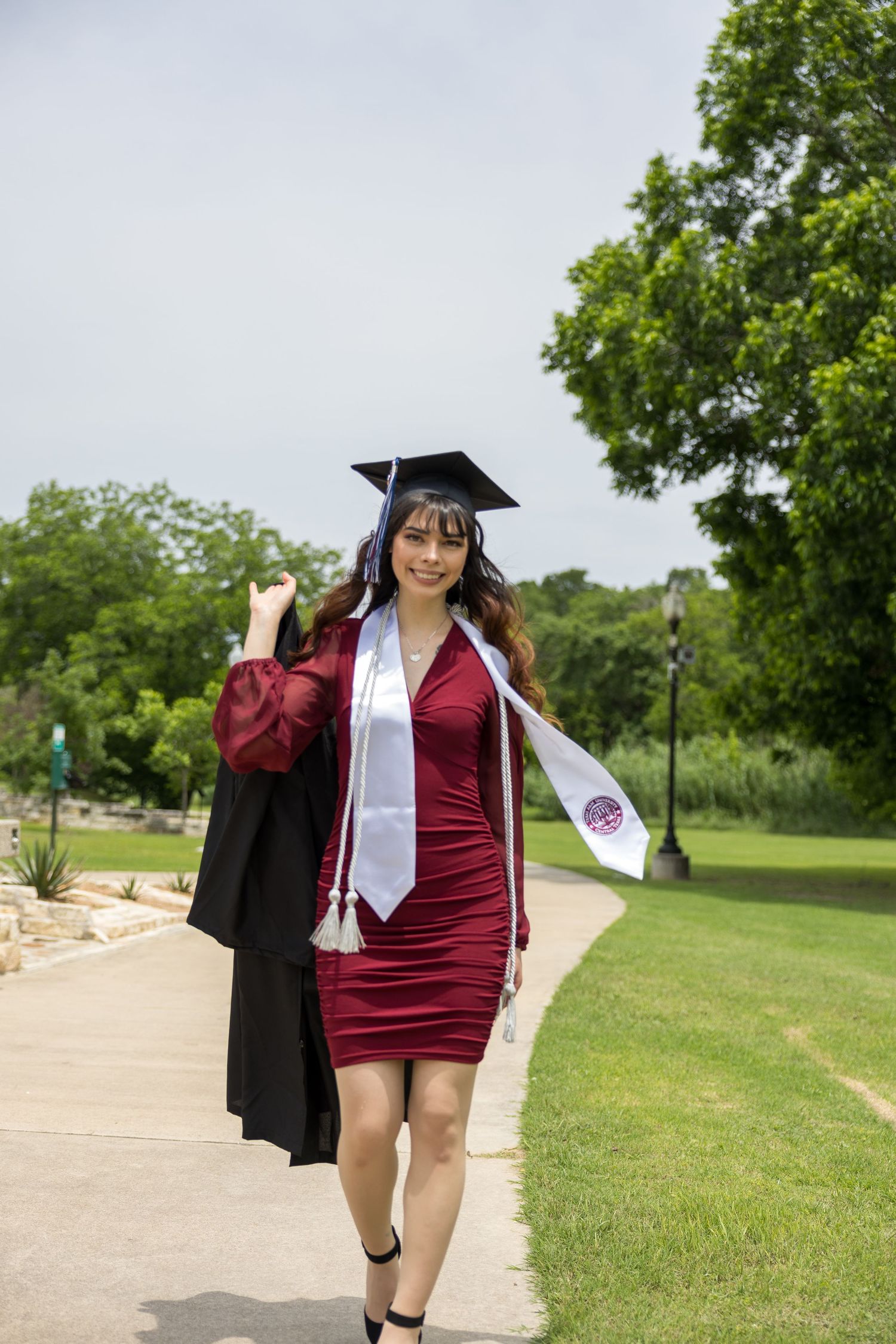 Graduate in burgundy dress and graduation cap poses in outdoor park setting with trees and pathway.