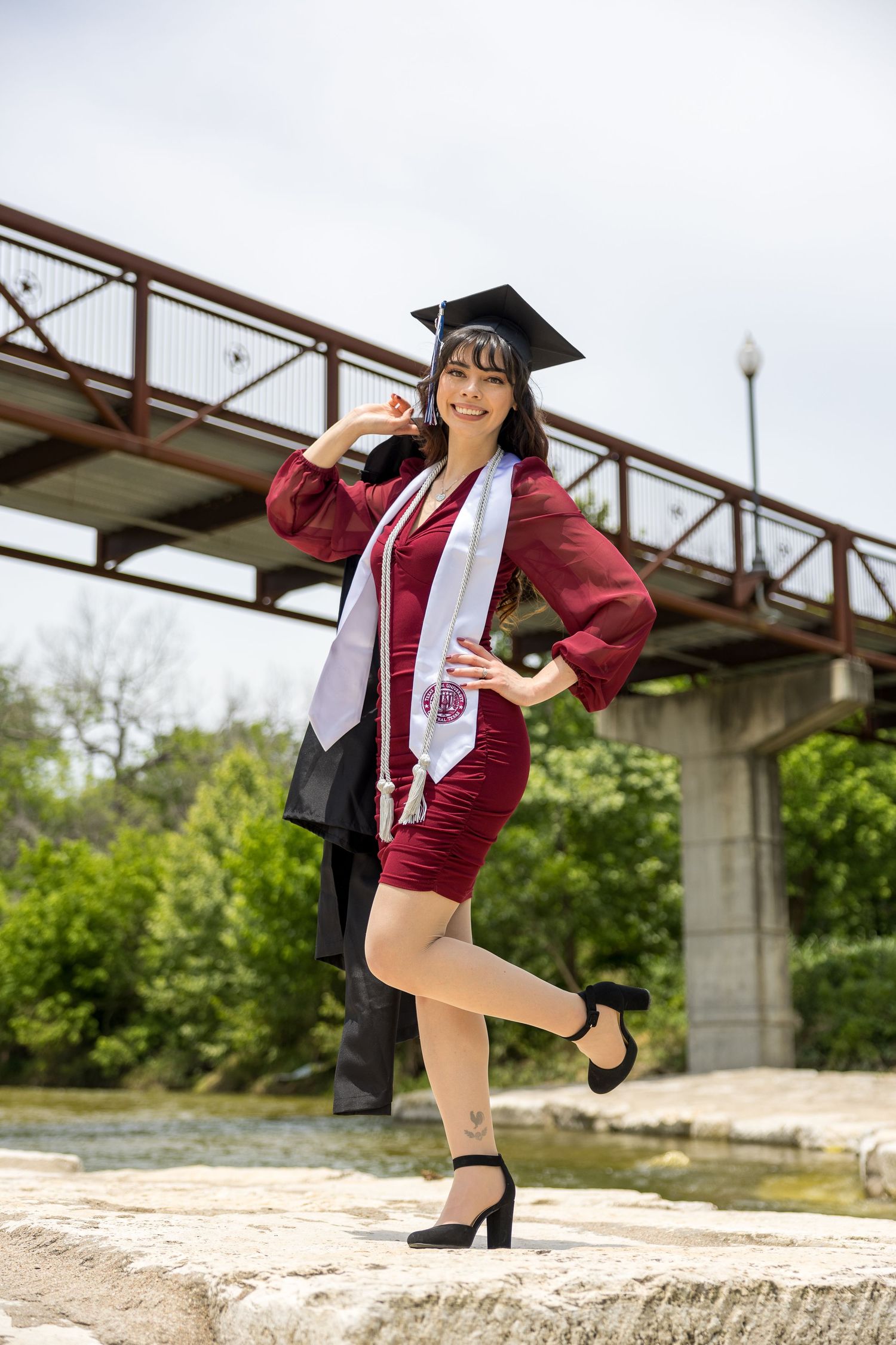 Graduate in maroon stole and black heels poses playfully near bridge structure during outdoor photo session.