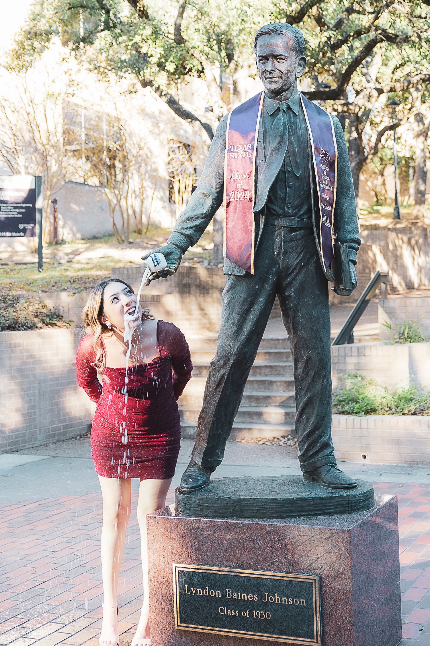 A playful interaction with a bronze statue of Lyndon Baines Johnson on a brick pathway surrounded by trees.