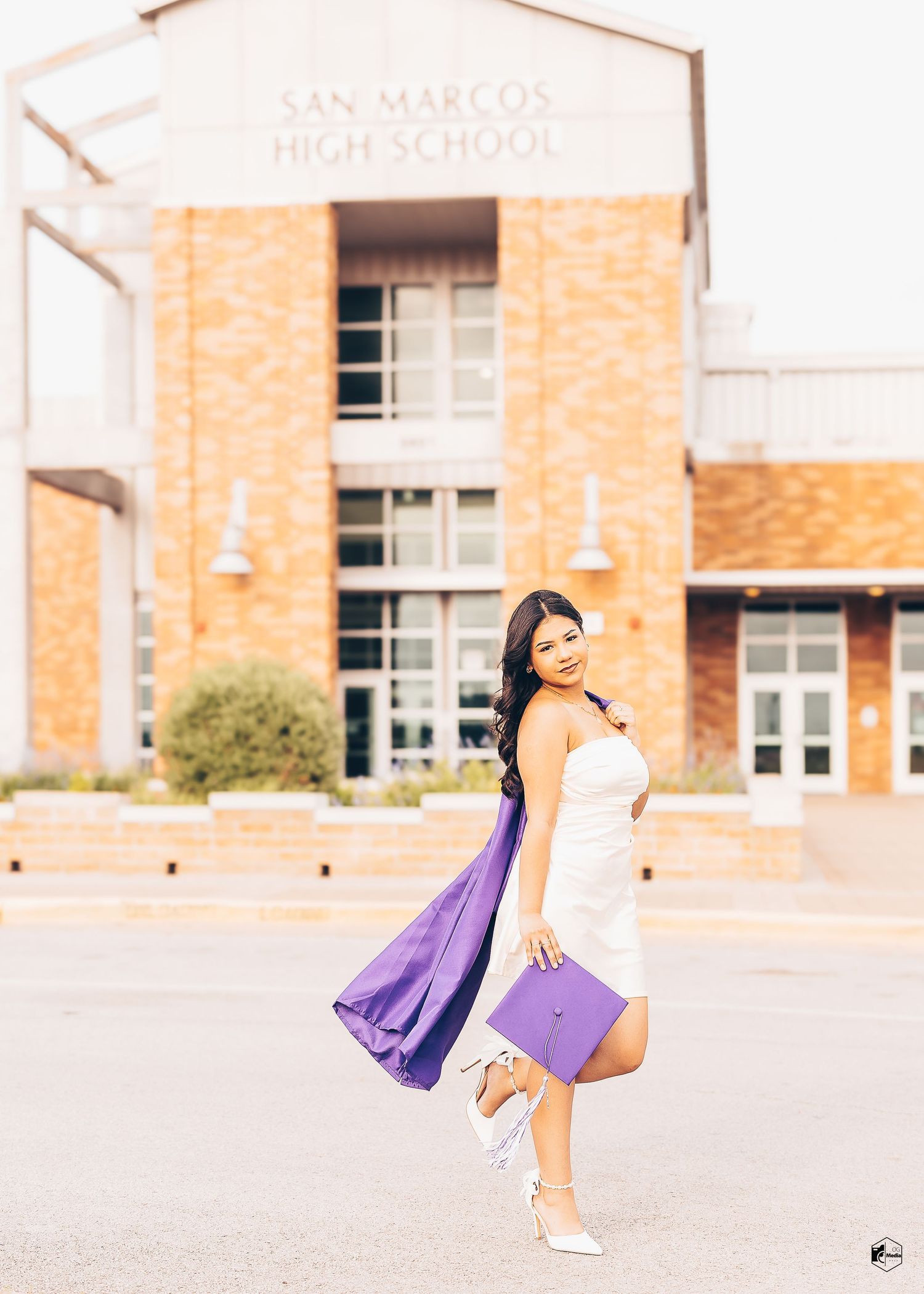 A person in a white dress and sneakers poses playfully in front of a brick school building on a sunny day.