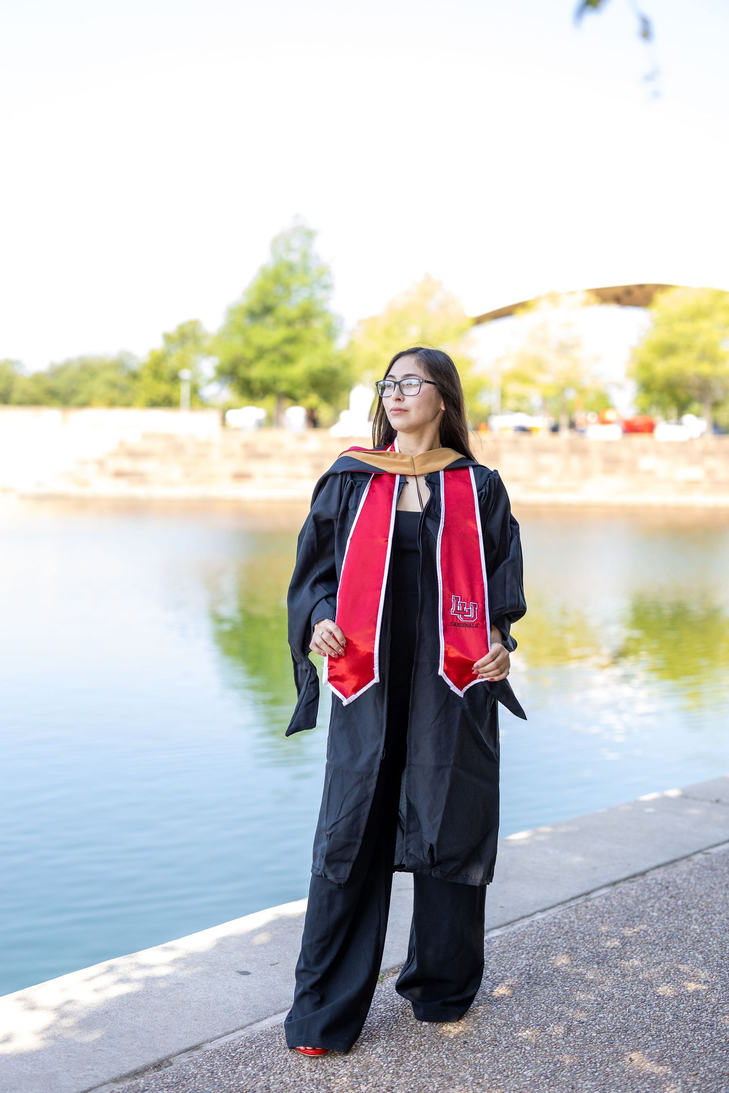 Graduate in black cap and gown with red stole poses by a lake on a sunny day for graduation photos.