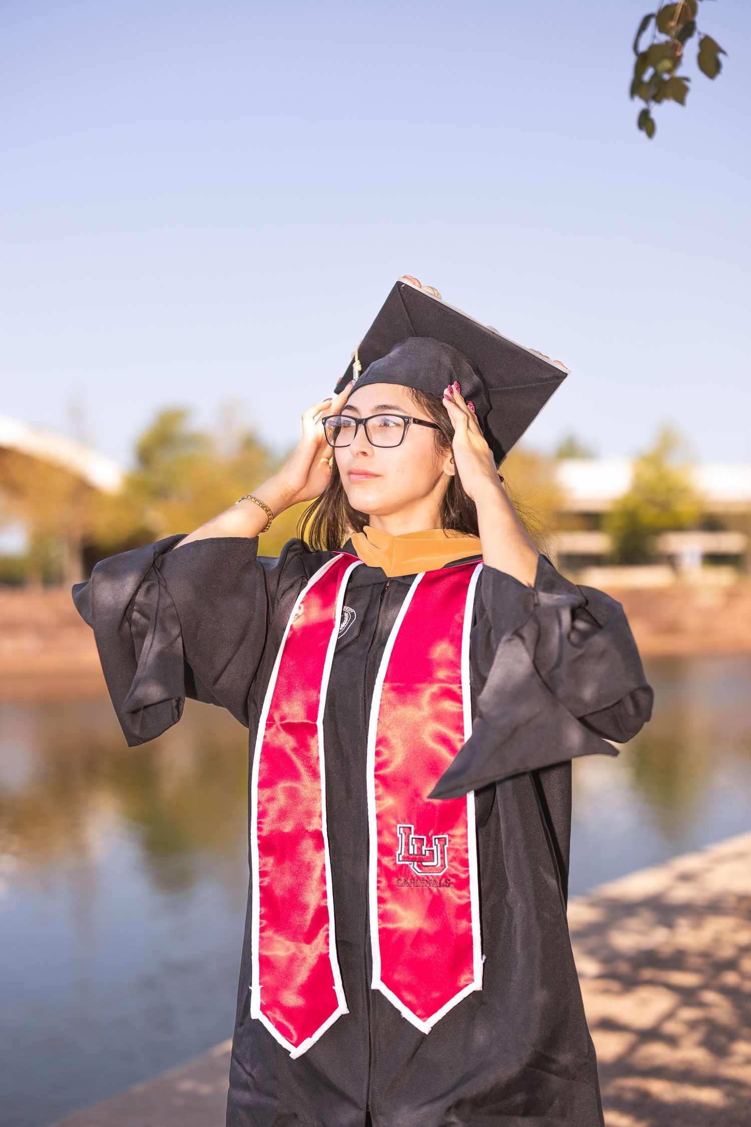Graduate in black cap and gown with red stole stands by lake during sunset celebration photoshoot.