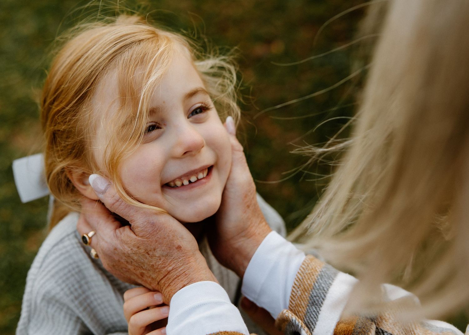 Loving grandmother gently holding smiling young girl with red hair by her cheeks outdoors.