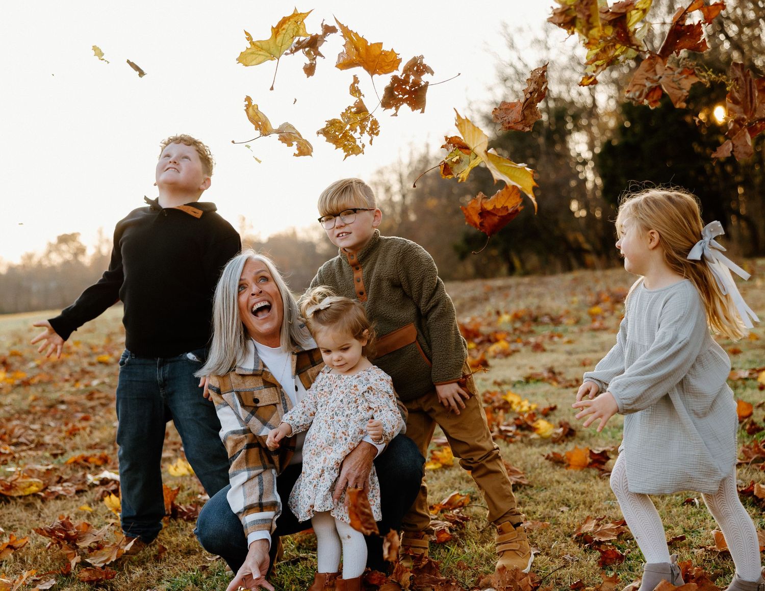 Joyful mother laughing with four children tossing autumn leaves in golden sunset light.