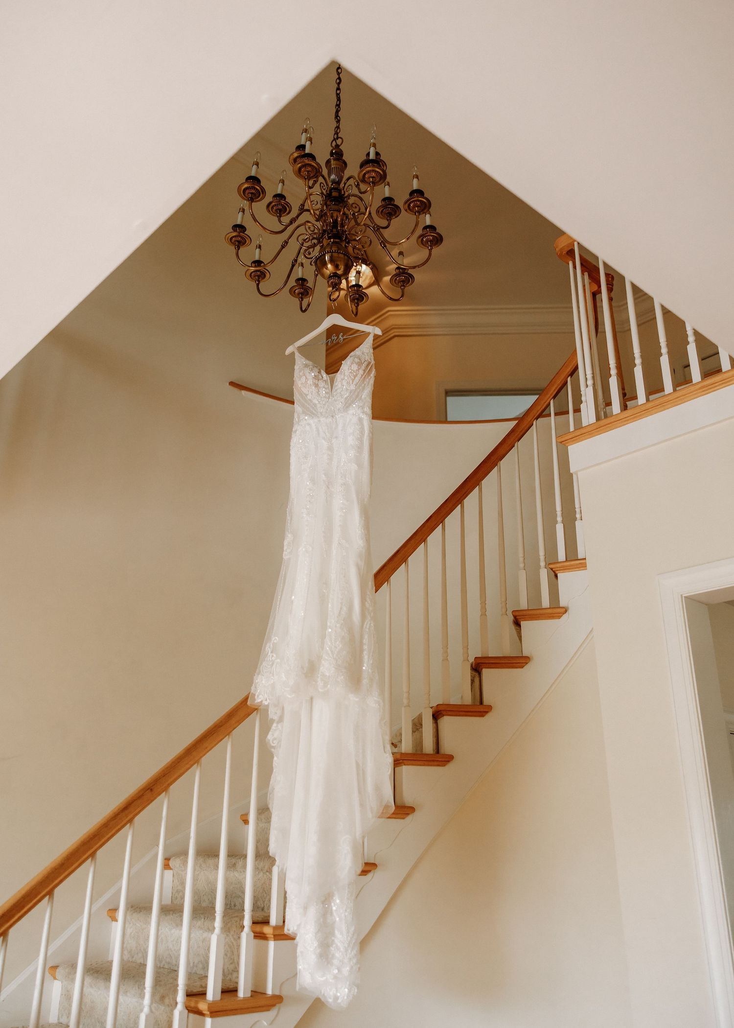 White lace wedding dress hanging from chandelier over elegant staircase with wood banister.